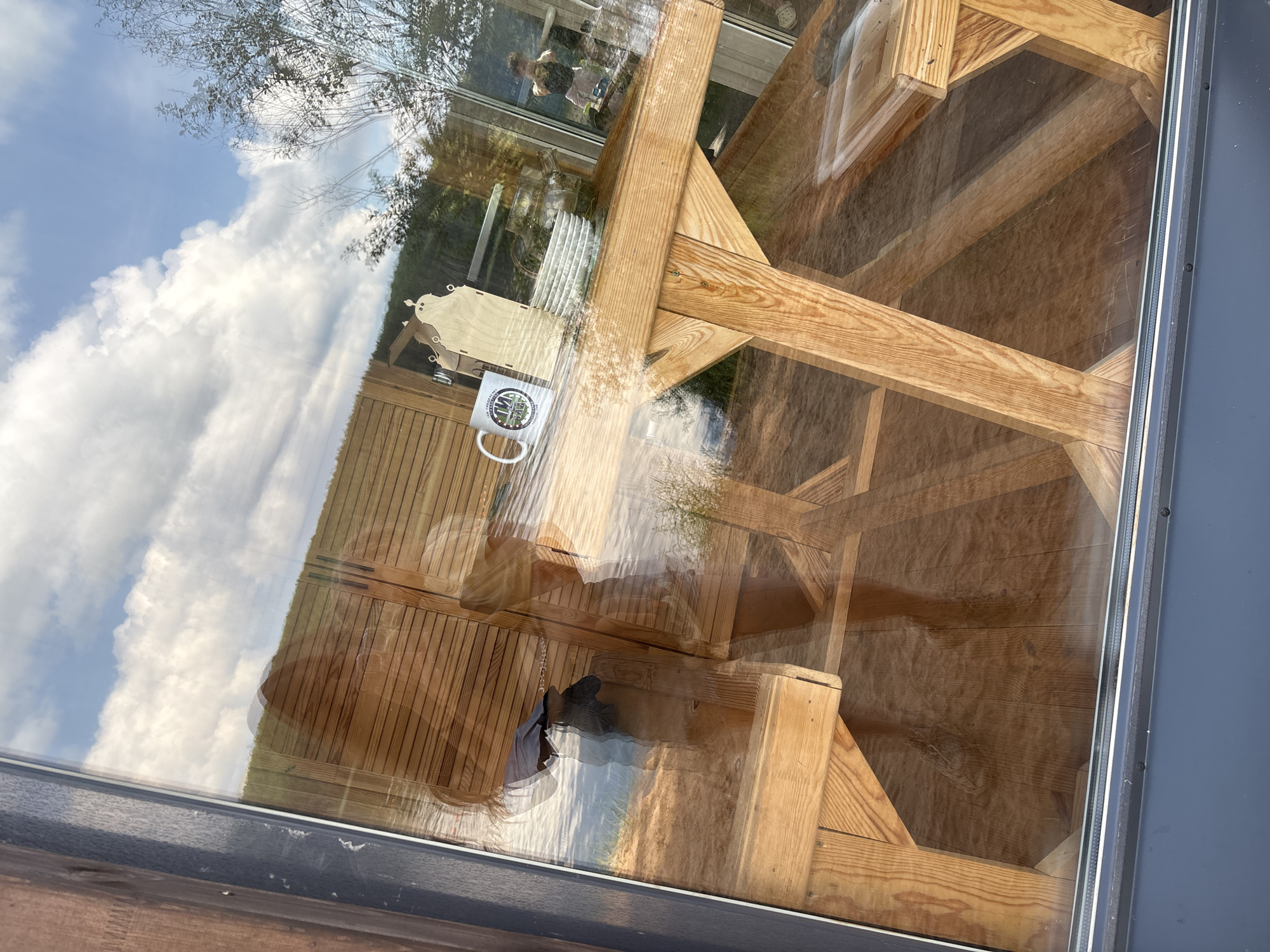This is a photograph taken through a window, showing reflections of the interior and the exterior scene. The exterior scene includes a bright blue sky with fluffy white clouds, trees, and a body of water, likely the Kama River, given the location in Chaykovsky, Russia. Reflected in the window is a wooden picnic table and benches. On the table, a white mug with a logo is visible. There are also reflections of people, including a person holding a phone and another person taking a selfie. The interior appears to be a room with wooden walls and flooring. The image captures a sunny day with a clear view of the sky and surrounding natural landscape.