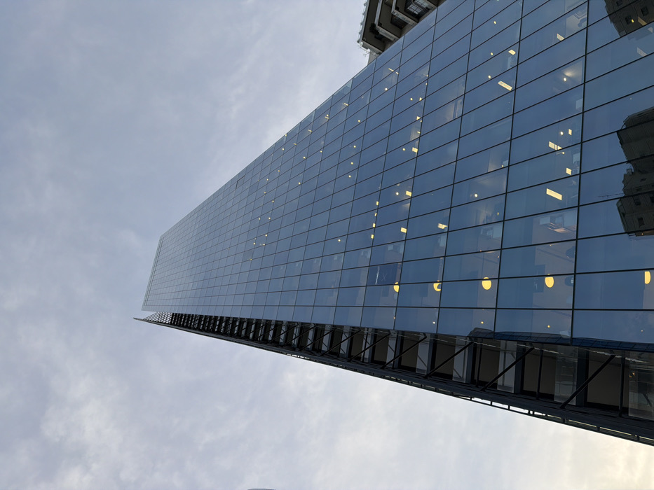 The image presents a tall, modern skyscraper in Ramat Gan, Israel, captured from a low-angle perspective looking upwards. The building's facade consists of a blue-tinted glass curtain wall arranged in a grid pattern. Internal lighting from various floors is visible through the glass, appearing as scattered yellow and white illuminated points and rectangular forms. Along the left edge of the building, a multi-tiered external structural element, composed of dark metal framing and transparent or dark panels, extends vertically. The upper section of the skyscraper tapers to a pointed architectural apex. The sky above is overcast or partially cloudy, exhibiting a uniform light gray hue. Reflections of a nearby, older architectural structure, characterized by stone or concrete elements, are discernible in the lower sections of the glass facade. No individuals, specific actions, or interactions are present within the scene.