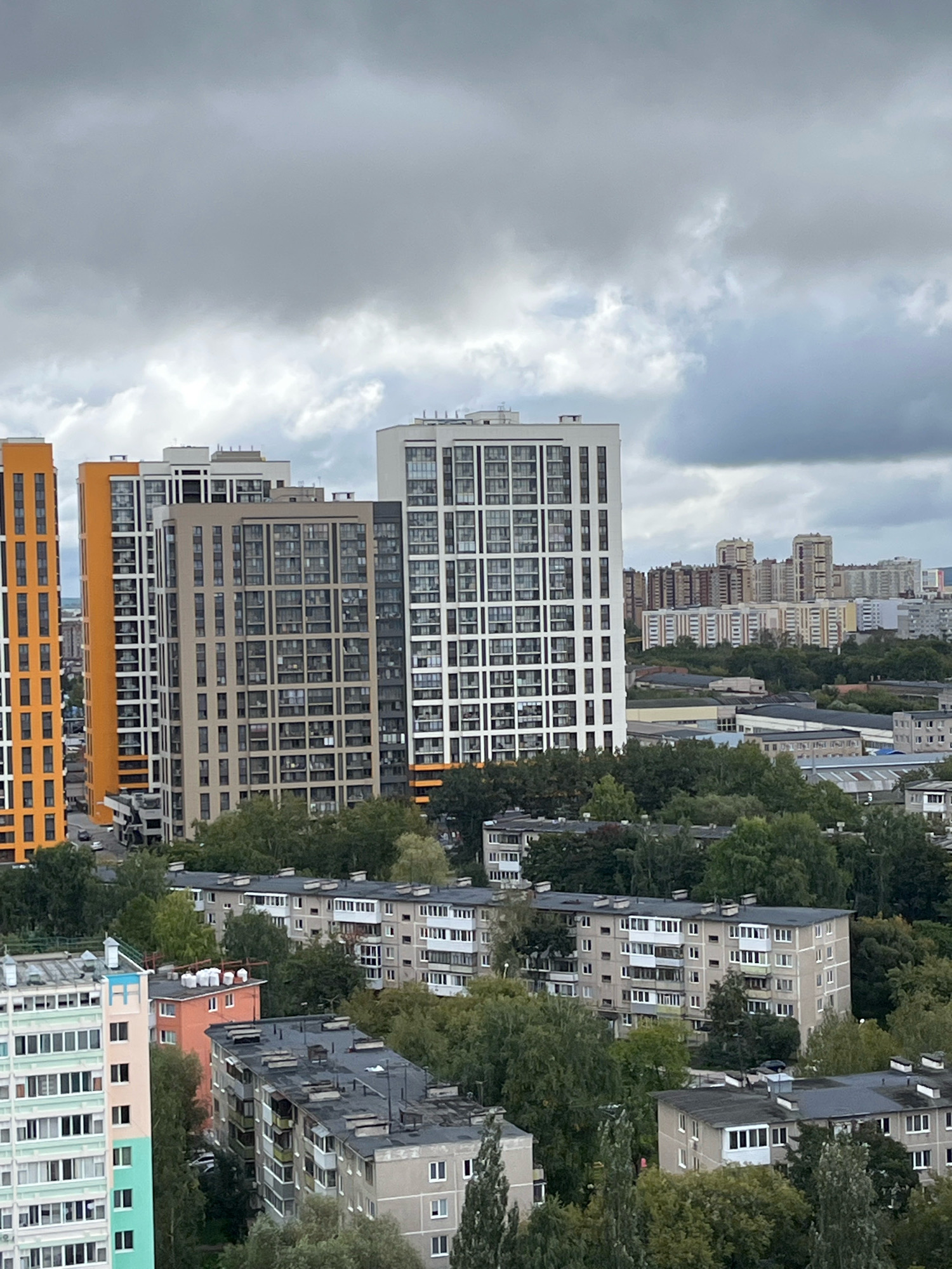The image depicts a panoramic view of a city neighborhood under a cloudy sky. In the foreground, several multi-story residential buildings are visible, characterized by their varied architectural styles and color palettes. Some buildings feature modern designs with large windows and balconies, while others appear older, with more traditional facades. Dense green trees intersperse the buildings, indicating a significant amount of greenery within the urban landscape.
In the mid-ground, more apartment blocks extend into the distance, some appearing as rows of identical units. Beyond these, a cluster of taller, more modern buildings rise, suggesting a developing or more central part of the city. The sky is heavily overcast with gray clouds, casting a diffused light over the entire scene. The overall impression is of a densely populated residential area, likely part of a larger urban development.
Based on the visual cues, such as the architectural styles of the buildings, the common use of balconies, and the presence of greenery, this scene is consistent with many residential areas in Perm, Russia. The mid-rise apartment blocks, interspersed with newer high-rises, are typical of the city's urban planning. The particular arrangement of buildings and the overall density suggest a specific district within Perm, possibly one undergoing modernization with the addition of new high-rise residential complexes adjacent to older Soviet-era housing estates.