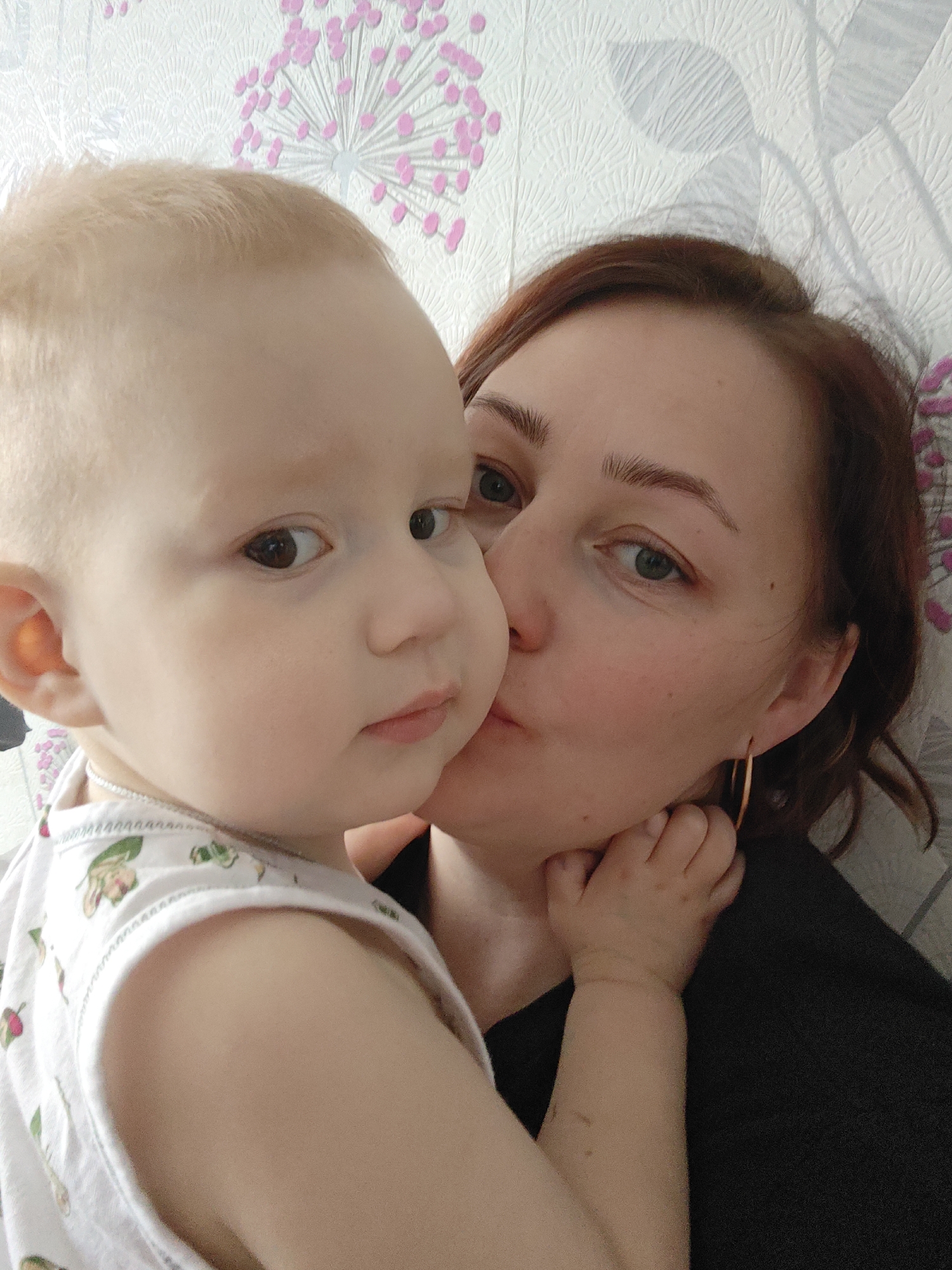 A fair-skinned adult female with reddish-brown hair is holding a fair-skinned baby with light blonde hair. The female is kissing the baby's cheek. The baby is wearing a white tank top with a floral pattern and a silver chain necklace. The female is wearing a black shirt and gold hoop earrings. They are positioned in front of a wall with a floral wallpaper pattern consisting of white lines and pink flower shapes. The image was taken in Markovskii, Russia.