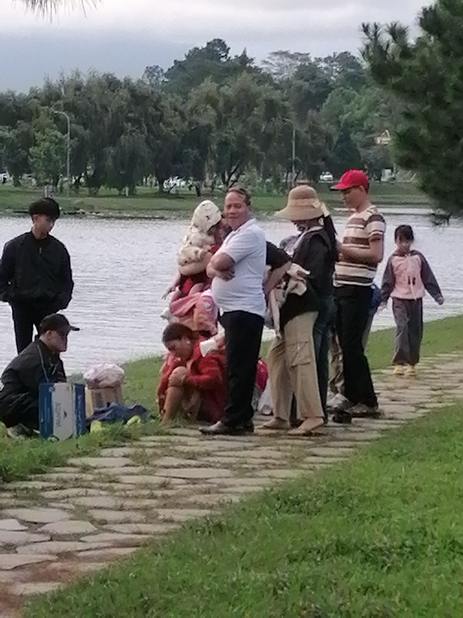 A group of people, including adults and children, are gathered on a paved pathway next to a body of water. In the foreground, a person in a black jacket and a cap is kneeling beside a blue and white box and a blue bag on the grass. Behind them, a person in a black jacket stands. To the right, a woman in a red jacket is crouched on the grass, and other individuals are standing and interacting. The background features trees and a body of water, suggesting an outdoor park or recreational area. The overall atmosphere appears casual and relaxed. The scene is likely located in Da Lat, Vietnam, known for its numerous lakes and parks.