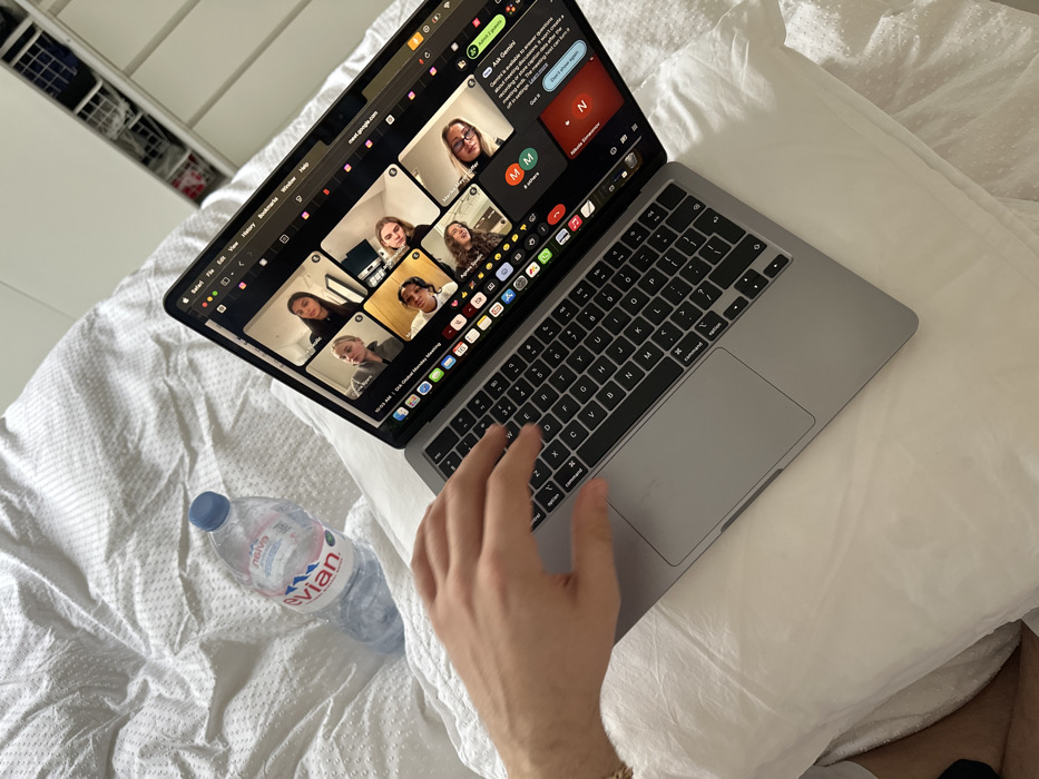 The scene depicts an indoor, residential setting in the city of London, United Kingdom. A silver Apple MacBook laptop is centrally positioned, resting on a white pillow atop a white, textured duvet. A person's right hand, adorned with a gold-colored bracelet, is positioned over the laptop's keyboard, indicating active use. To the left of the laptop, a clear plastic Evian water bottle with a blue cap is visible on the duvet. The laptop screen displays an active Google Meet video conference, timed at 10:03 AM and titled "DFA Global Monday Meeting." The conference features a grid view of at least seven participants, predominantly young women, with names such as "Agnes G." and "Marnie W." visible. A chat sidebar on the screen shows a message from "Nikos Kliamouri" and an "Ask Gemini" AI prompt. In the upper right background, a white shelving unit containing various items is partially visible.