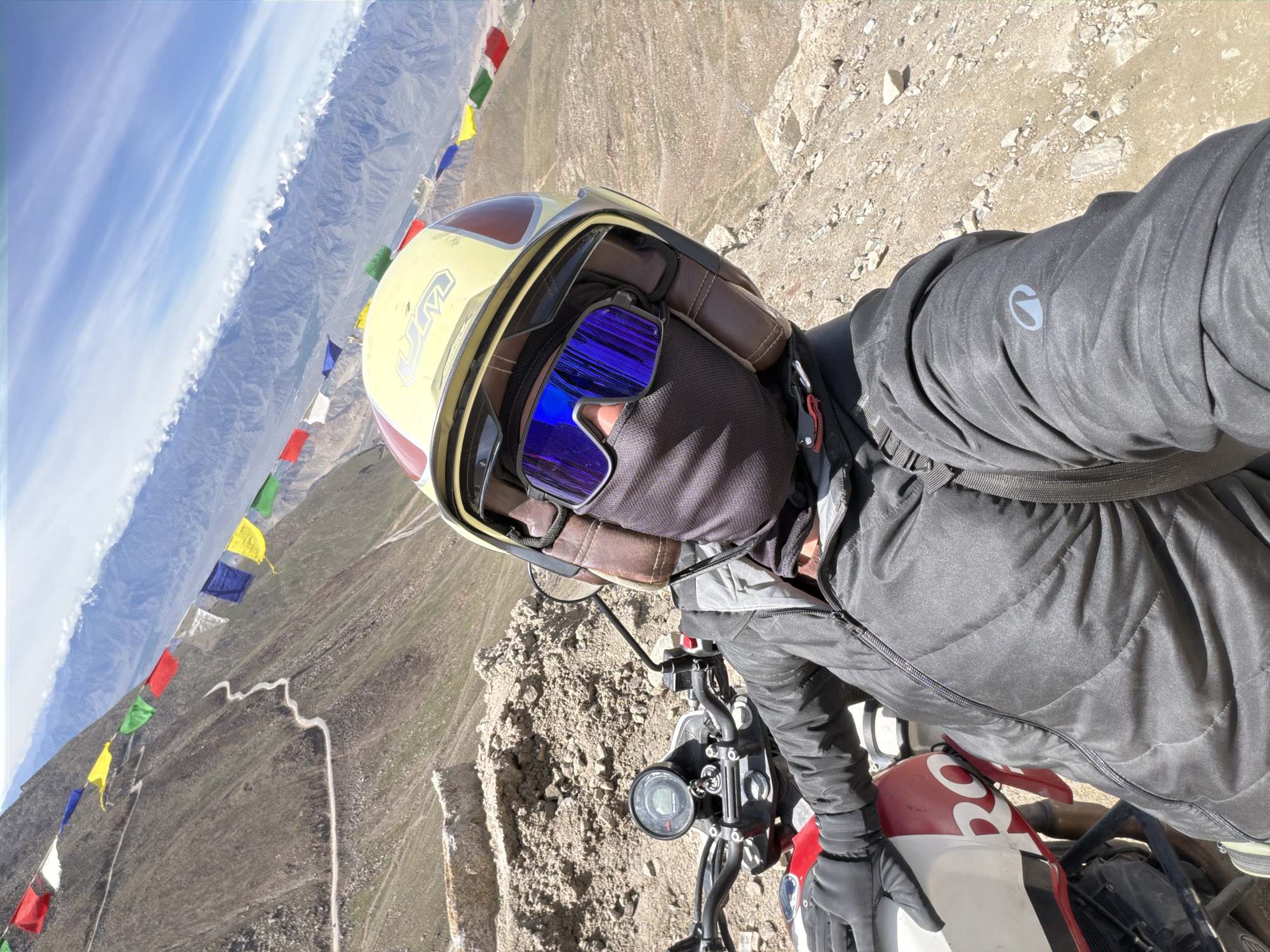 A first-person perspective selfie is captured on a motorcycle in Leh, India, during the daytime. The subject, wearing a yellow and red helmet with brown leather trim, blue mirrored goggles, and a dark face mask, is seated on a motorcycle. The motorcycle's dashboard shows a speedometer and other indicators. The subject is dressed in a black puffer jacket, black gloves, and a backpack. The background reveals a steep, mountainous landscape with a winding road and colorful prayer flags strung across the mountainside. The sky is blue with scattered clouds. The terrain is rocky and arid, typical of the Himalayan region around Leh.
