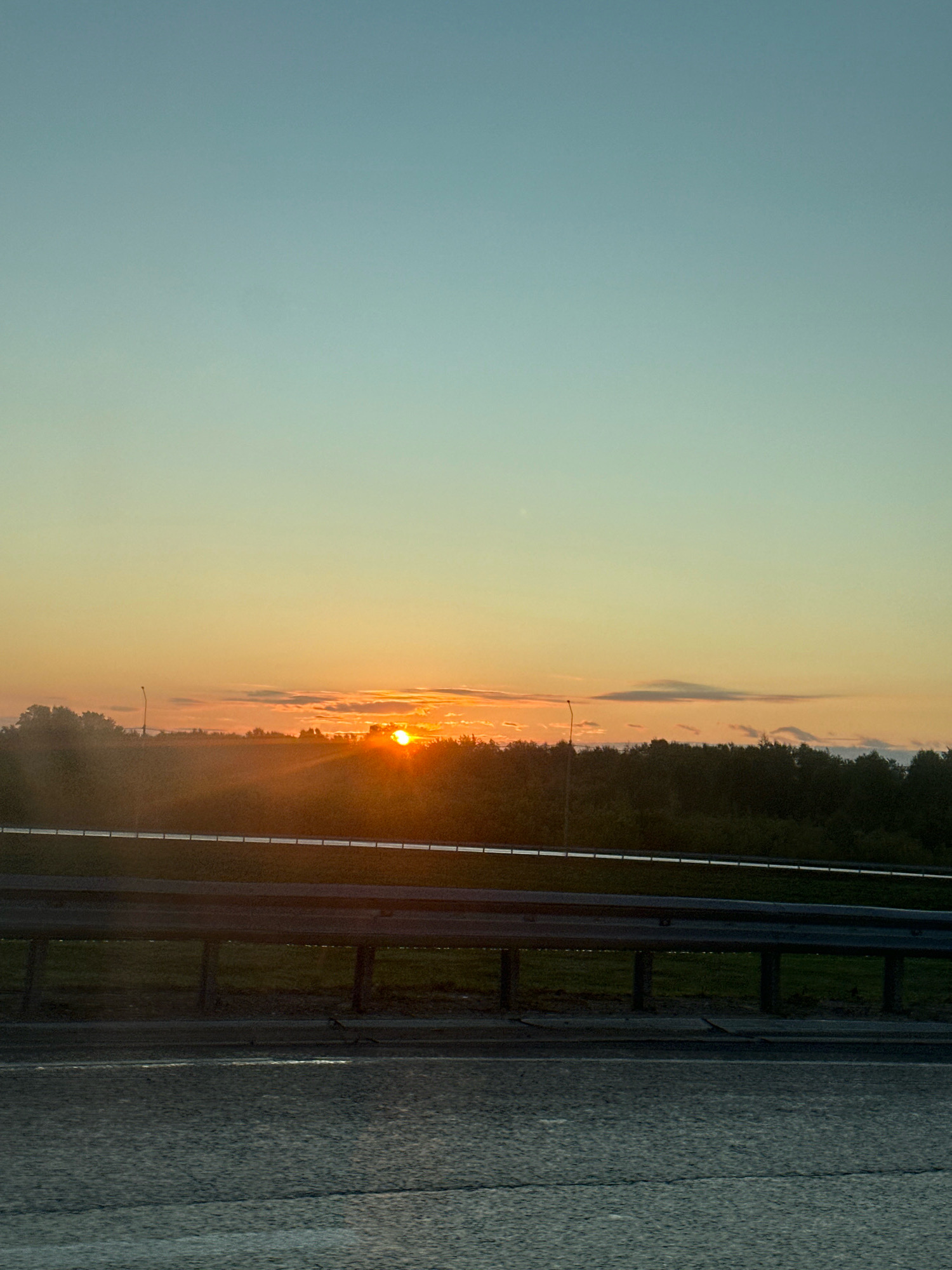 An outdoor, eye-level, medium shot captures a sunset over a highway. The sun, a bright orange orb, sits low on the horizon, partially obscured by faint clouds and peeking through a line of dark green trees. The sky transitions from a pale yellow-orange near the sun to a soft blue at the top of the frame. A gray metal guardrail runs horizontally across the middle ground of the image, separating the highway from a grassy embankment. The asphalt of the highway is visible in the foreground, showing faint white lines. The overall lighting is warm and diffused, characteristic of a sunset. The location is not discernible from the image.