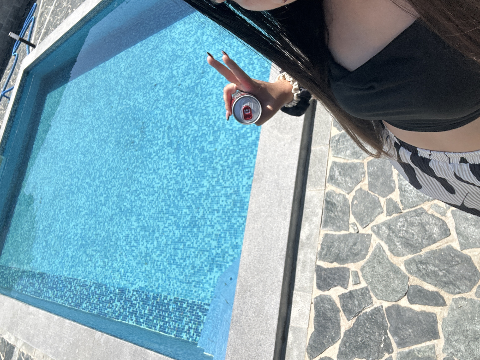 The image provides an overhead, angled view of a person by a swimming pool on a bright, sunny day. The person's upper body is visible, with long dark hair cascading down their right side. They are wearing a black crop top and black and white striped pants. Their right hand is holding an open red and white beverage can, with two fingers (index and middle finger) extended in a gesture that resembles a peace sign or "V" sign. A white scrunchie is visible on their right wrist, and their fingernails are long and dark.
The primary setting is a swimming pool filled with clear, blue water. The pool's interior is lined with small, square mosaic tiles in shades of blue, and a darker band of blue tiles runs along the bottom section of the visible pool wall. A light gray concrete coping surrounds the edge of the pool. Adjacent to the pool and coping is a stone patio, characterized by irregularly shaped gray stones set in lighter-colored grout. In the top left corner, a blue rail or pipe is partially visible next to the pool.
The bright lighting indicates it is daytime, likely a sunny morning or afternoon. The scene suggests a leisure activity or relaxation by the poolside. No discernible text is visible on the beverage can or elsewhere in the image.