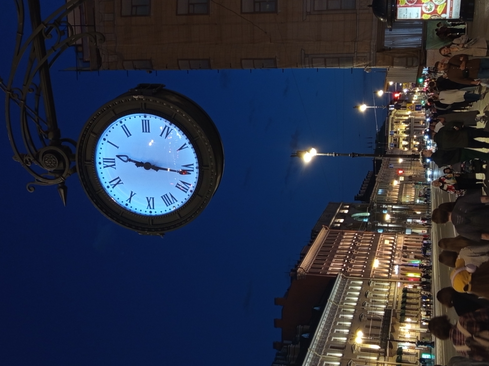 A large, illuminated clock face with Roman numerals hangs over a street at dusk in Saint Petersburg, Russia. The clock, supported by an ornate metal bracket attached to a building on the right, displays the time as approximately 10:08. The street is lined with illuminated buildings displaying classical architecture, with lights on in the windows and storefronts. A moderate number of pedestrians are visible on the sidewalks and crossing the street. Traffic lights and street lamps illuminate the scene. The sky is a deep blue, indicating nighttime.