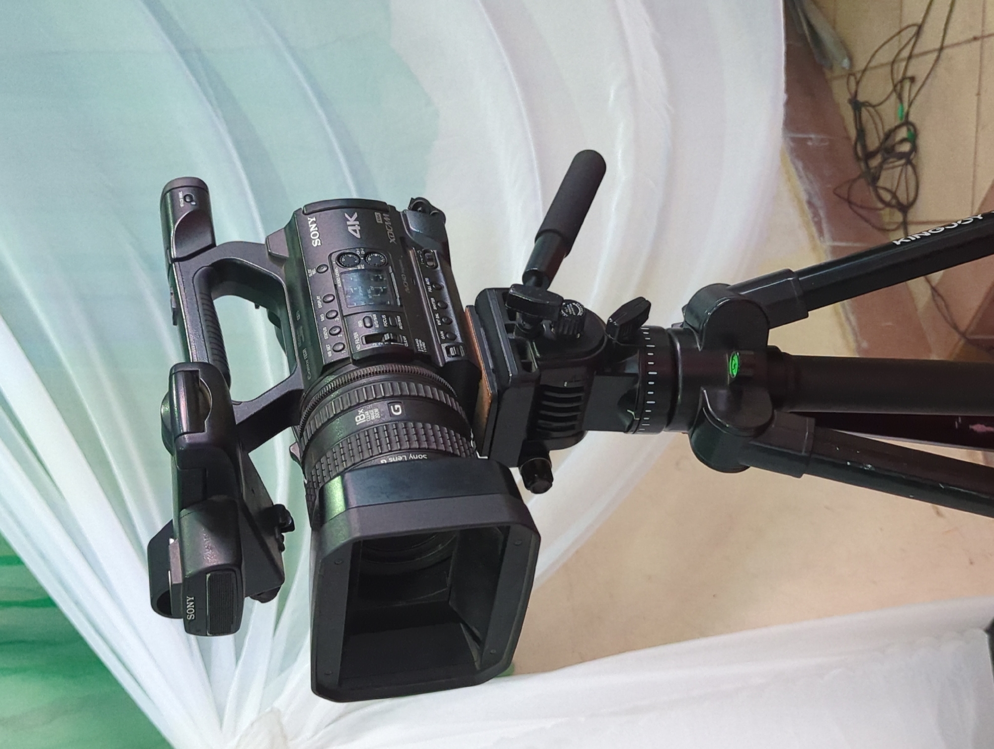 A detailed shot shows a Sony 4K video camera mounted on a black Kingjoy tripod. The camera is pointed to the left, showing its lens, controls, and grip. It is positioned in front of a white fabric backdrop, possibly used for video or photography. The environment appears to be indoors, but specific location details are not provided.