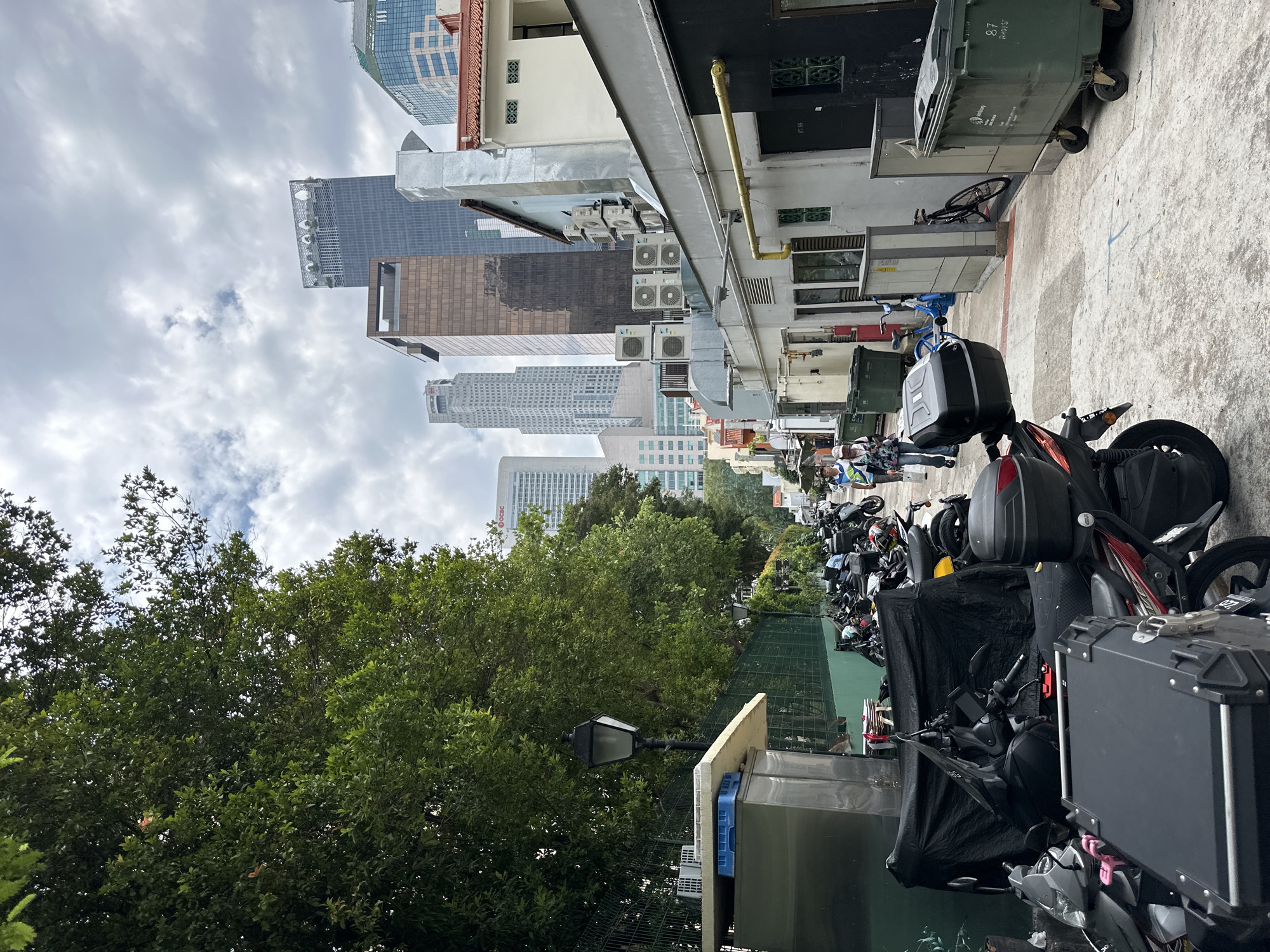 This is an eye-level, outdoor, medium shot of a narrow alleyway in Singapore. In the foreground on the left, several motorcycles are parked, some are covered with a black tarp. One motorcycle has a large black storage box on the back. In the middle ground, a person in a white helmet and blue shirt is walking away from the camera. On the right side of the alley, there are buildings with air conditioning units and trash bins. In the background, modern skyscrapers loom over the scene. The sky is cloudy.