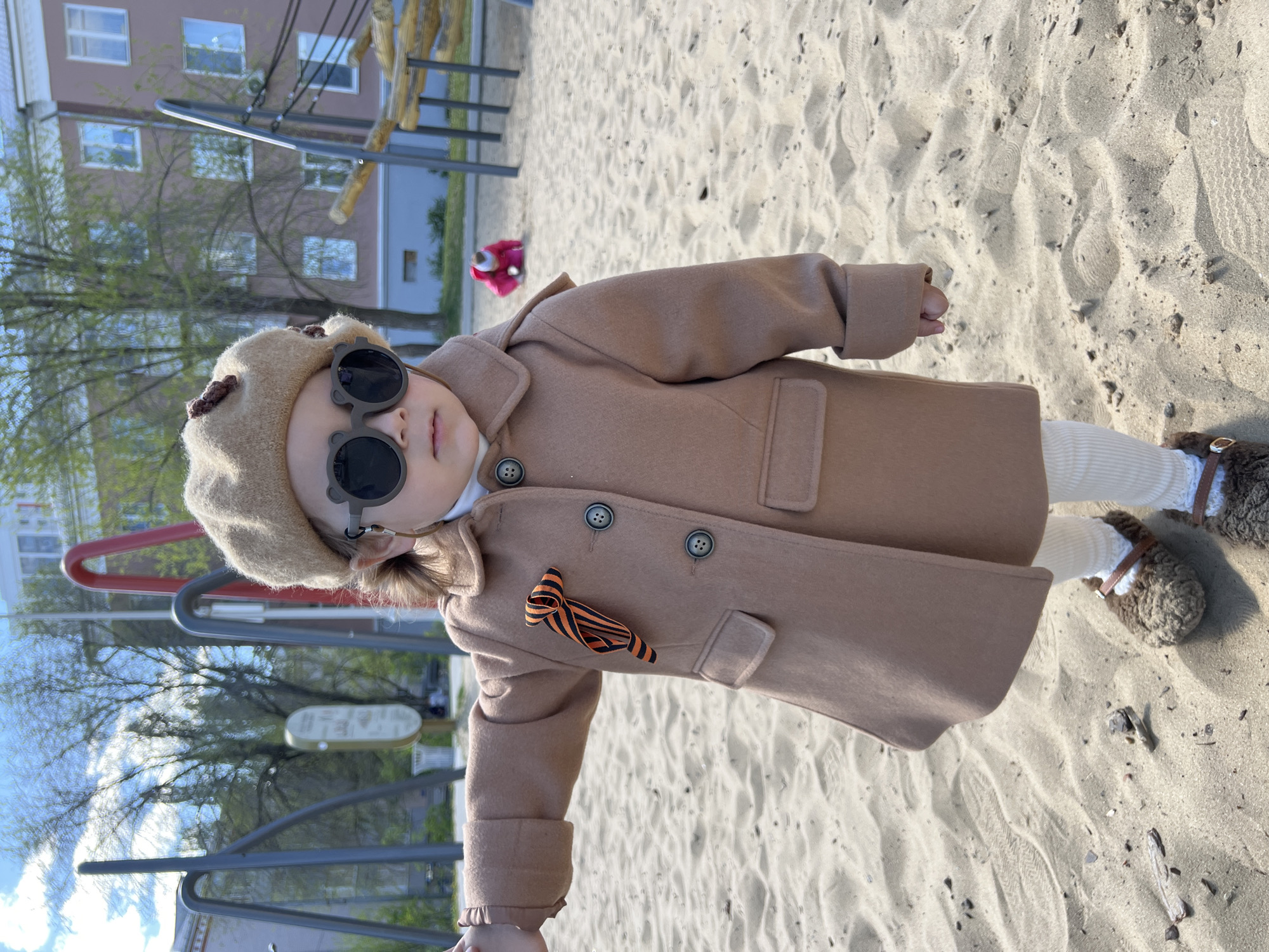 A young child stands in a sandbox, wearing a tan coat, matching beret, and round sunglasses. A black and orange ribbon is pinned to the coat. The child is also wearing white tights and brown shoes. The background shows playground equipment, a building, and trees. The location is Kazan, Russia.
