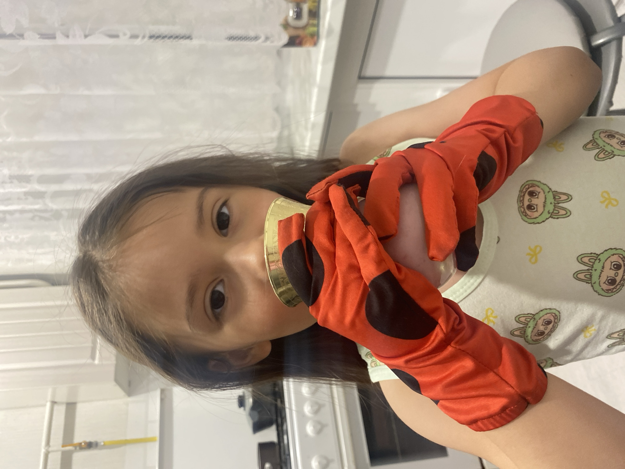 The image, taken in Chaikovsky, Russia, features a young girl engaged in playful activity indoors. She is positioned facing the camera, with her head tilted, and her dark hair falls around her shoulders. Her face is partially obscured by a golden, reflective object, possibly a cup or a toy, which she holds up to her mouth or nose area.
The most striking elements of her attire are a pair of bright red gloves with black polka dots, reminiscent of a Ladybug costume, worn on both hands. She wears a light-colored garment, possibly pajamas or a t-shirt, adorned with repeating cartoon patterns of green bunny faces and small yellow bow motifs.
The setting appears to be an indoor space, likely a kitchen or a brightly lit room within a home. The background shows white or light-colored horizontal blinds or curtains, and what look like white kitchen cabinets or wall panels. Hints of household appliances, possibly a stove or oven, are visible in the blurred background. A light-colored chair or stool is also partially visible on the right. The lighting suggests an indoor setting, possibly with artificial light, making the time of day ambiguous. The girl's visible eye shows a focused or curious gaze as she engages with the golden object. No discernible text is visible in the image.