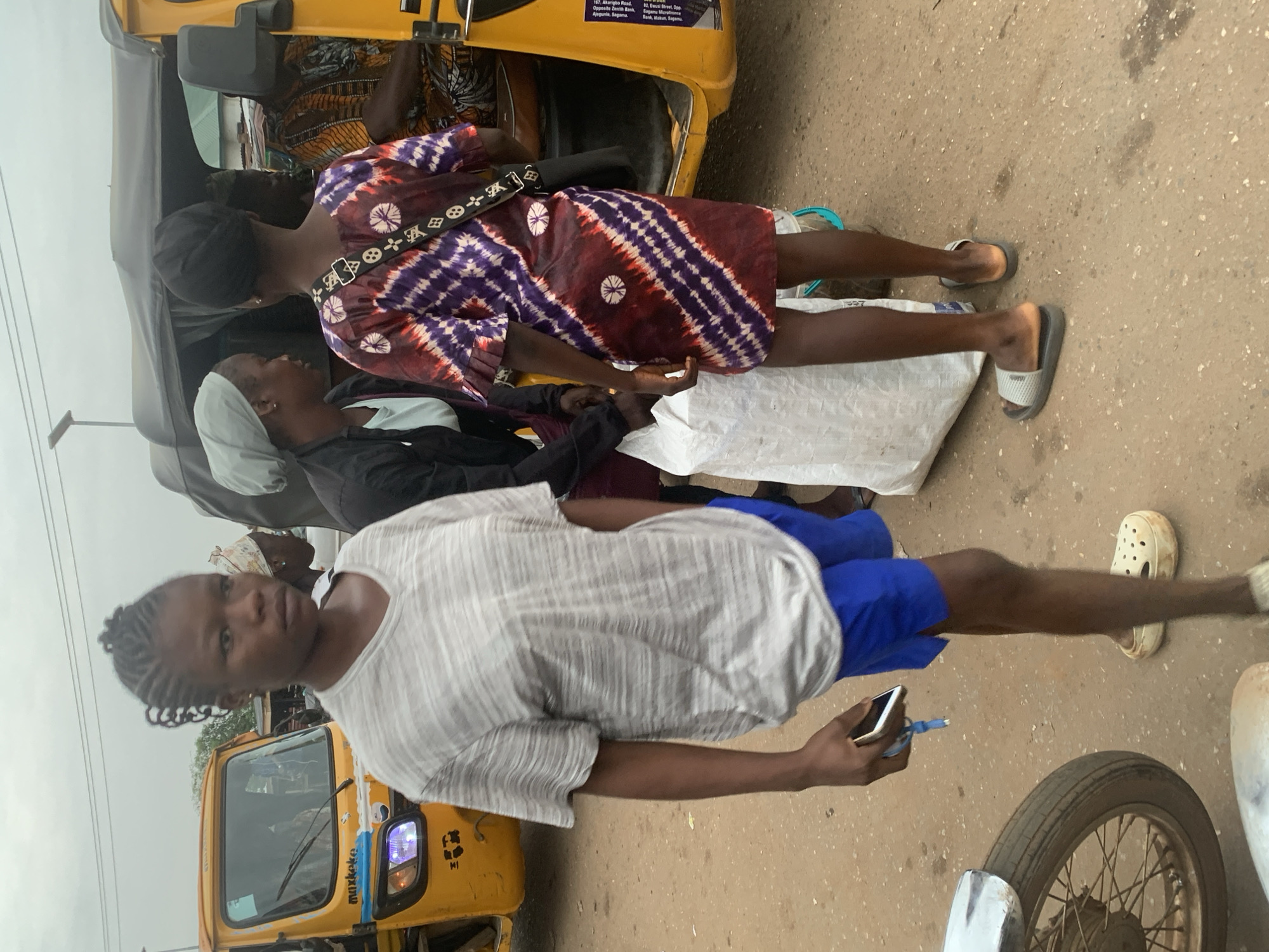 The image depicts a street scene in Shagamu, Nigeria, likely a market or transportation hub given the presence of a yellow auto-rickshaw (also known as a Keke Napep) and other individuals.
In the foreground, a young woman with braided hair pulled back stands facing left, looking slightly off-camera. She is wearing a grey, horizontally striped t-shirt and blue shorts. Her right hand holds a smartphone with a blue charging cable attached. She is wearing white clog-style sandals.
Behind her, to her right, another individual, likely a woman, is facing away from the camera, wearing a black headscarf and a dark jacket over a lighter top. She is standing next to a large, white sack.
Further to the right, partially obscured by the sack and the auto-rickshaw, is a person wearing a colorful, tie-dye patterned dress and a black headscarf. A black strap with white symbols, resembling a designer logo, is slung across their shoulder. This person is standing near the open door of the auto-rickshaw.
The auto-rickshaw is yellow with a grey canopy. The name "Muxkeke" is visible on the side of the vehicle. The ground is dirt or unpaved road, with scattered debris. In the far left of the image, a portion of a motorcycle wheel and fender is visible. The background shows other people and structures, suggesting a bustling urban environment under an overcast sky. The overall lighting is diffused and appears to be daytime.