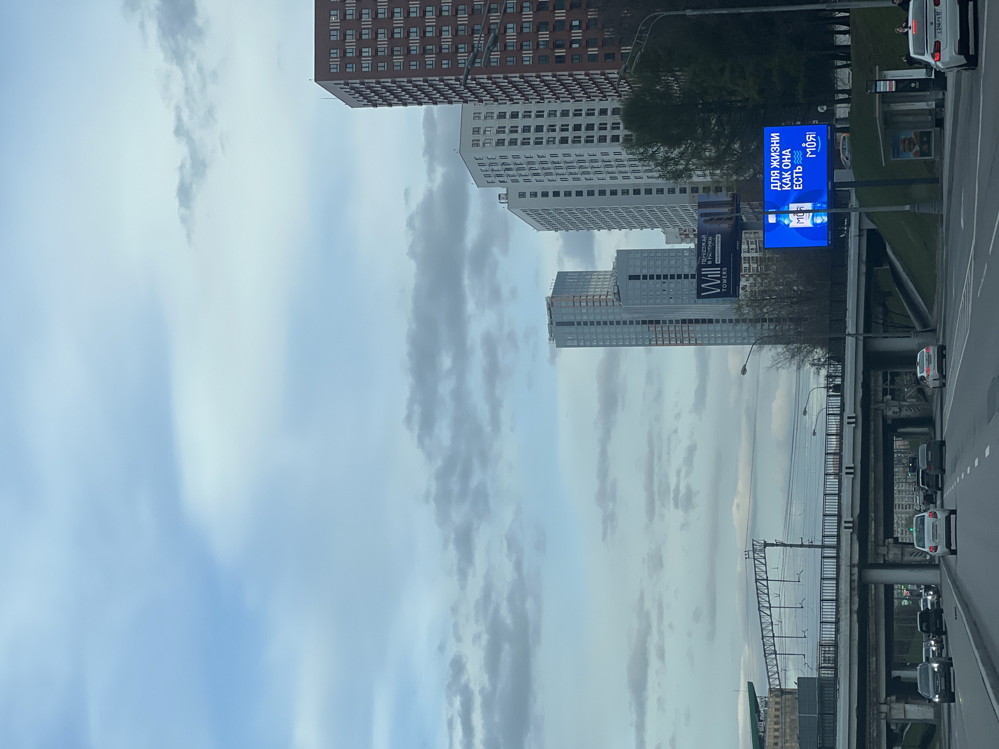 The image shows a view from a bridge in Khimki, Russia, featuring a row of tall buildings against a cloudy sky. On the right side of the image is a busy roadway with traffic. A large, blue billboard displaying an advertisement in Russian is visible near the roadway. Power lines and other infrastructure are also present.