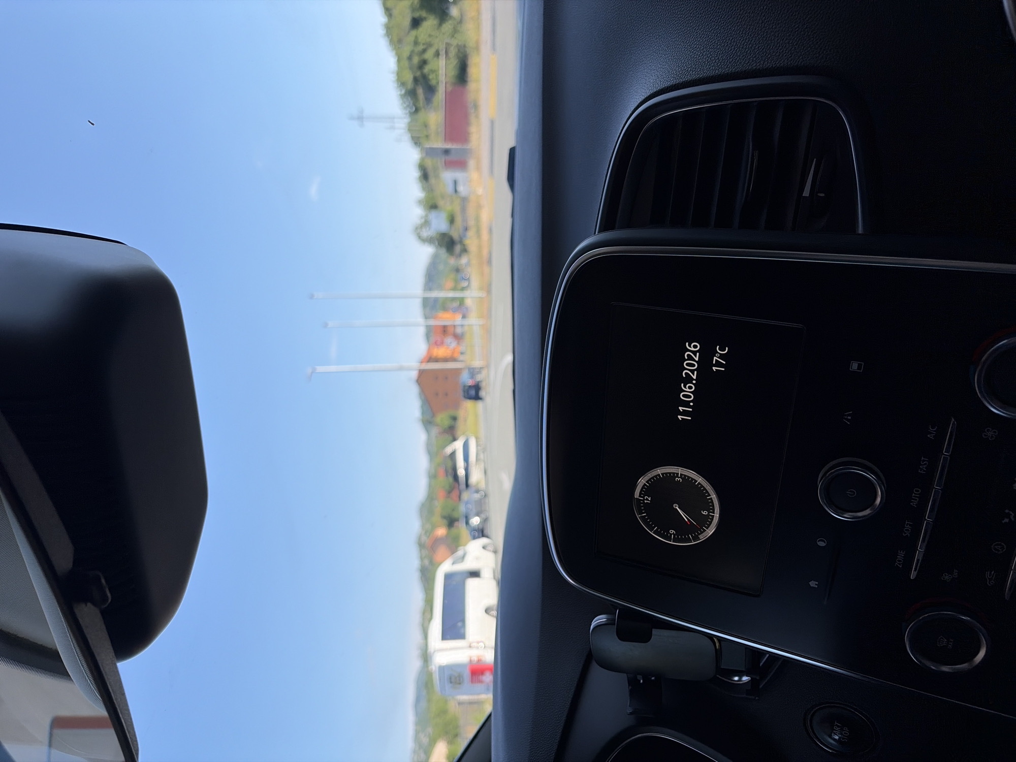 The image, taken from the interior of a car, shows a dashboard display with a clock and the date 11.06.2026, along with a temperature reading of 17°C. Outside the car, through the windshield, a street scene unfolds under a clear blue sky. Several vehicles are visible, including a white van with red markings, and other cars. Buildings and hills with vegetation form the background. The scene appears to be in Očinići, Montenegro, based on the context provided.