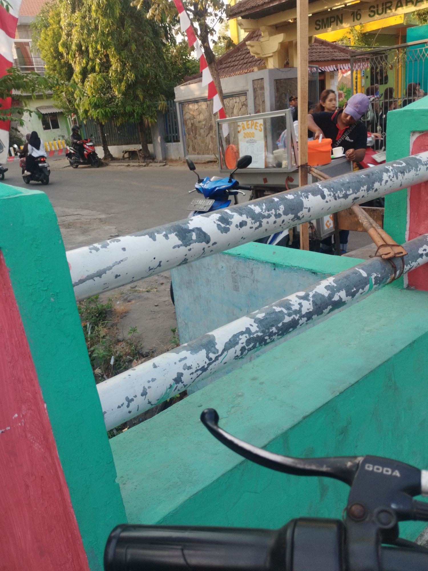 In front of SMPN 16 Surabaya, a crepes stand is visible with a vendor preparing food. Several people are gathered around the stand, and a few motorcycles are parked nearby. The scene depicts a typical street-level view of a school area in Surabaya, Indonesia, with everyday activities taking place. Red and green painted concrete barriers and metal railings are in the foreground.