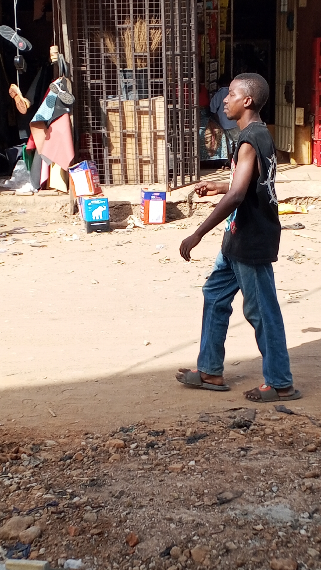 A young man with short hair wearing a black sleeveless shirt and blue jeans walks along a dirt and gravel road in Sabon Tasha, Nigeria. He is wearing sandals. To the left, a shop displays various items including shoes, clothes, and boxed goods. A metal grate and wooden pallets are also visible. The surrounding area appears dusty and exposed.
