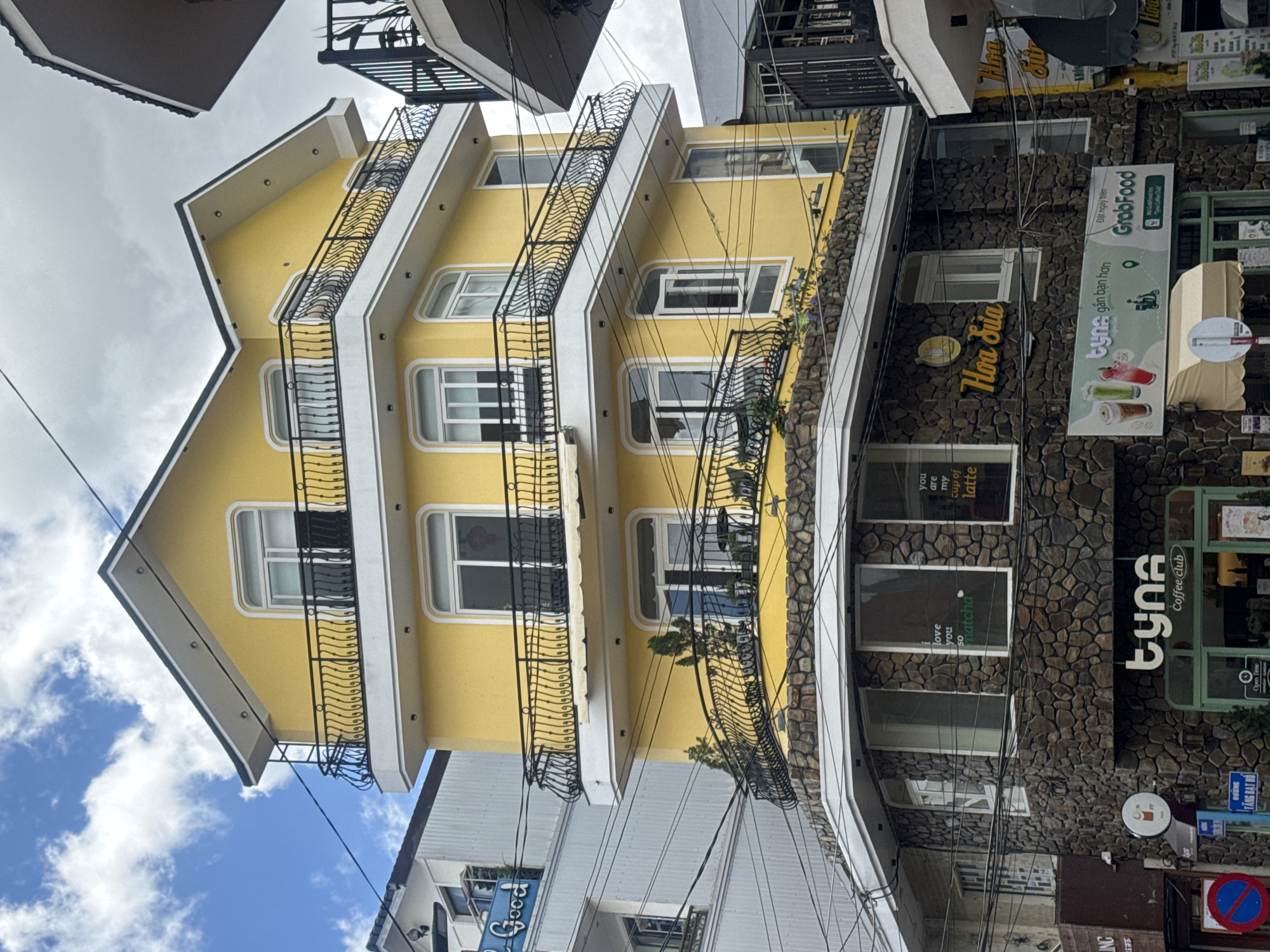 The image shows a bright yellow building with multiple balconies and ornate black railings in Da Lat, Vietnam. The building has a stone-clad lower level with several windows. Signs for "tyna Coffee Club" and "Hoa Sua" are visible on the lower facade, as well as a GrabFood advertisement. Various power lines crisscross in front of the building. The sky is partly cloudy.