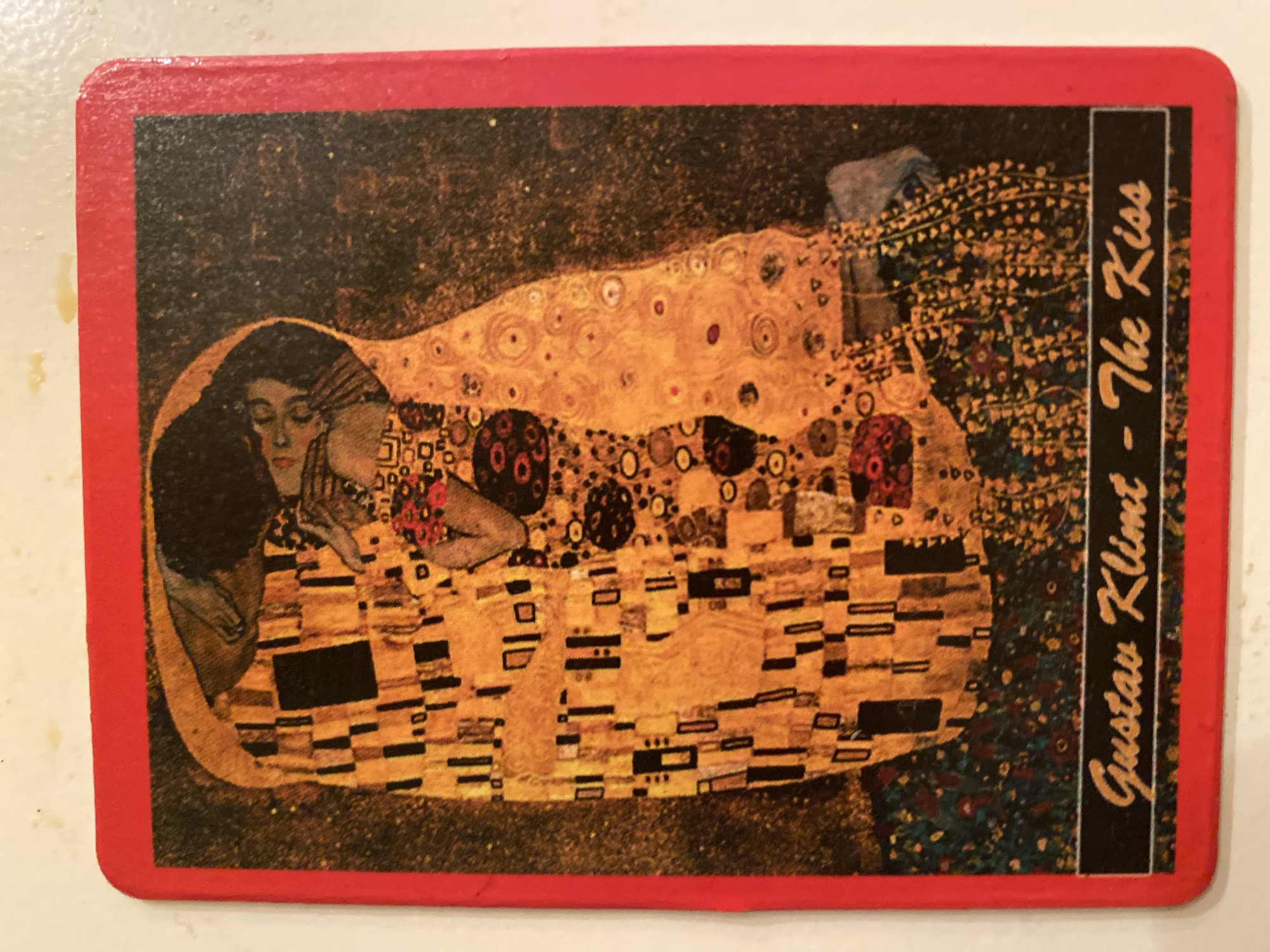 The image shows a rectangular postcard featuring Gustav Klimt's painting "The Kiss." The postcard has a red border. The painting depicts a couple embracing, rendered in Klimt's signature Art Nouveau style with opulent use of gold leaf and decorative patterns. The male figure, clad in a robe adorned with rectangular shapes, is shown kissing the female figure, who is kneeling and wearing a richly patterned dress. The background is dark and textured, with scattered golden and circular motifs. Below the artwork, a black rectangular band contains the text "Gustav Klimt - The Kiss" in a stylized script. The postcard is lying on a light-colored surface, possibly a table, with some minor imperfections visible on the surface. The location in the city of Ivanteyevka, Russia is not discernible from the provided image.