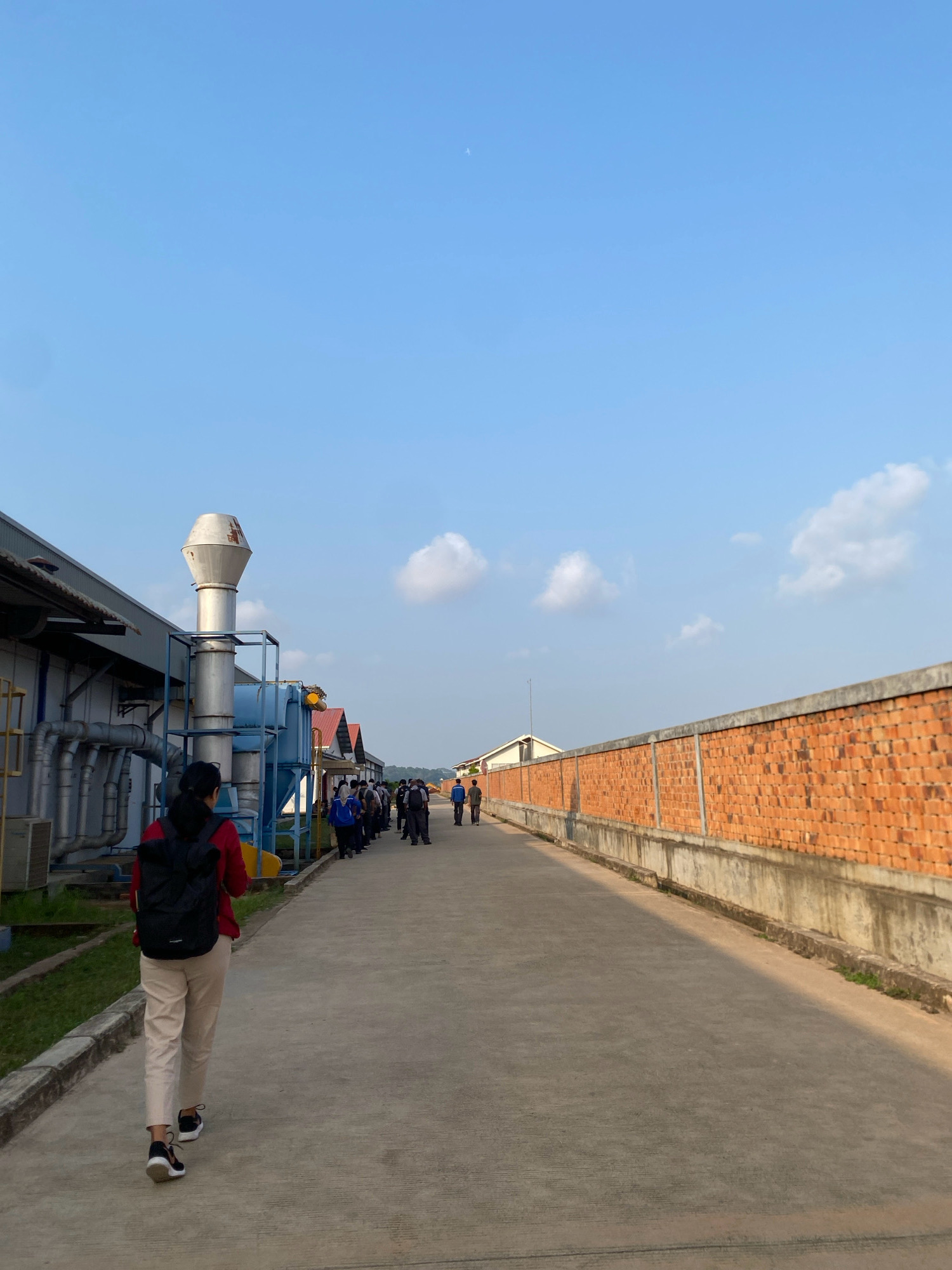 A woman with a black backpack and a red shirt walks away from the camera on a concrete path. She is wearing light-colored pants and black and white sneakers. To her left is a building with industrial piping and a large ventilation stack. To her right is a brick wall with concrete support structures. In the distance, a group of people are walking in the same direction as the woman. The sky is clear with a few clouds and a faint airplane contrail. The scene appears to be an industrial area in Batam, Indonesia.
