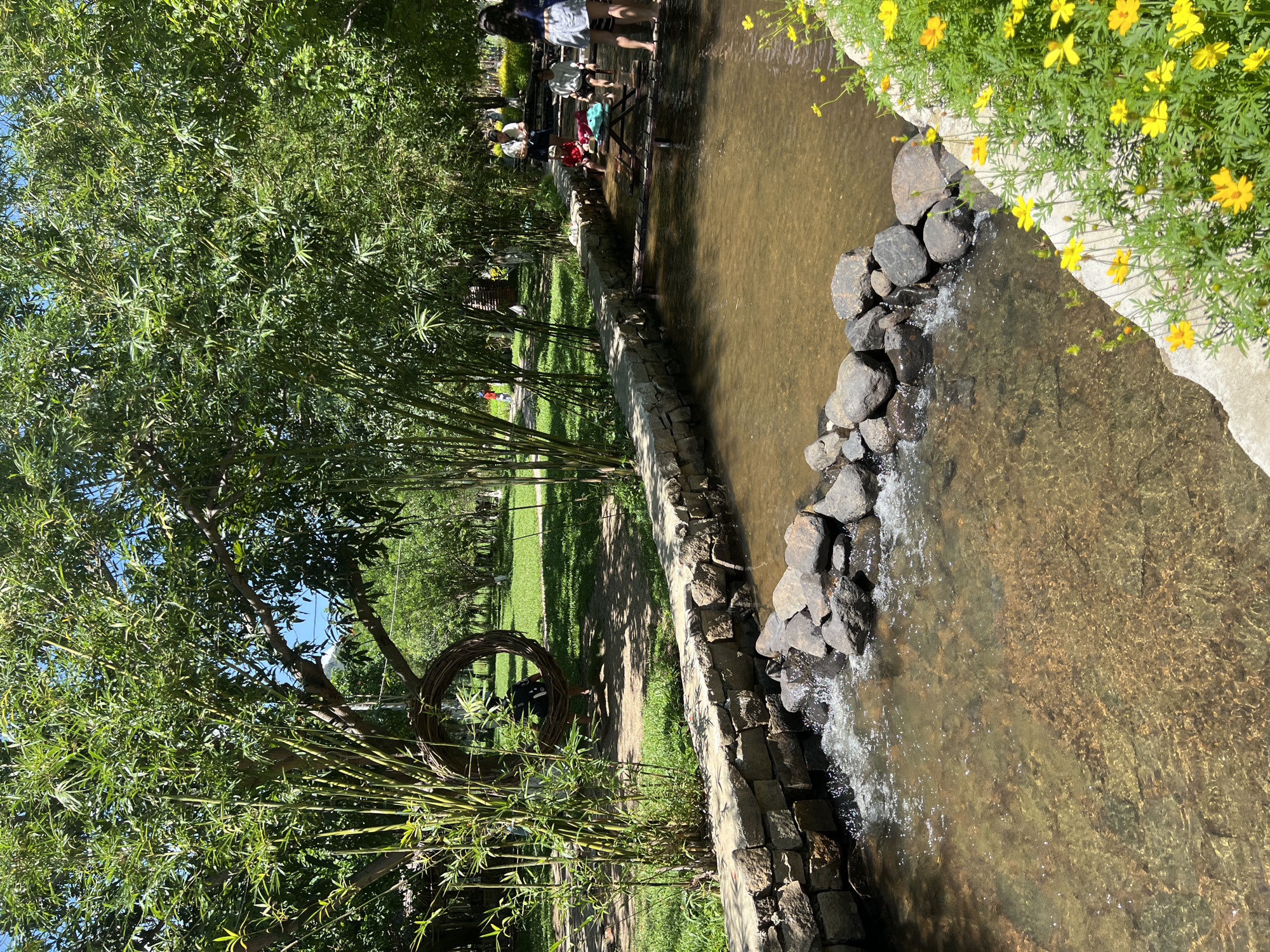 The image depicts a serene outdoor scene, likely in Nam Cam Ranh, Vietnam, featuring a shallow, clear stream flowing over a bed of rocks and gravel. The stream is bordered by a low, constructed wall of rough-hewn stones. Lush green trees and bamboo stalks flank the stream, creating a shaded, natural ambiance. In the background, several people are visible, some sitting near the water's edge and on a wooden structure extending into the stream, while others stand or walk on the grassy bank. One individual is seated in a circular woven swing hanging from a tree branch. Yellow flowers are blooming along the edge of the stream in the foreground. The overall impression is a tranquil recreational area in a verdant environment.