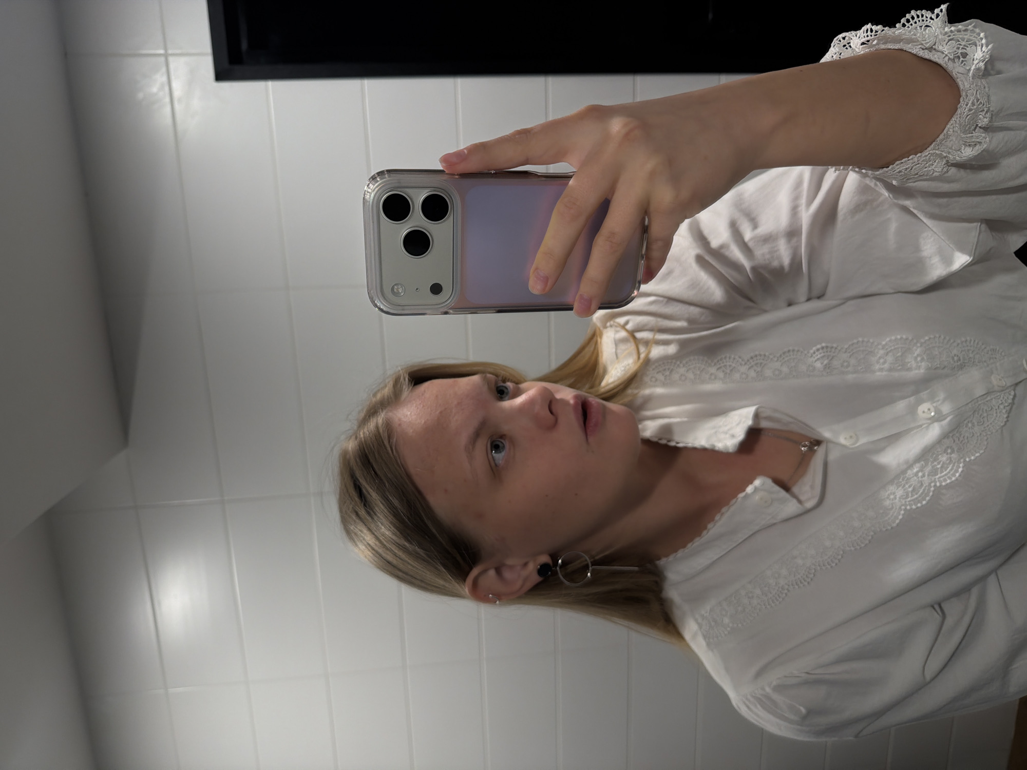 This is an indoor mirror selfie taken in Izhevsk, Russia, featuring a young woman. The image is rotated, presenting the woman's figure horizontally across the frame. She has blonde hair and is wearing a white long-sleeved blouse adorned with delicate lace patterns on the collar, front placket, and ruffled cuffs. Her left ear is visible, featuring two small silver stud earrings and a larger, distinct black and silver circular or bar-shaped earring. A thin silver necklace hangs around her neck.
The woman is holding a light-colored smartphone, equipped with a clear case and a distinctive triple-camera array, in her right hand. The phone is angled to capture her reflection in what appears to be a mirror. Her gaze is directed slightly upwards, likely at the phone screen, and her mouth is slightly open, indicating an expression of focus or momentary surprise during the selfie-taking process.
The background consists of white tiled walls, suggesting an interior space like a bathroom. A dark, rectangular object or frame is visible in the upper left portion of the image, potentially the top edge of a mirror or a cabinet. No discernible text is visible in the image.