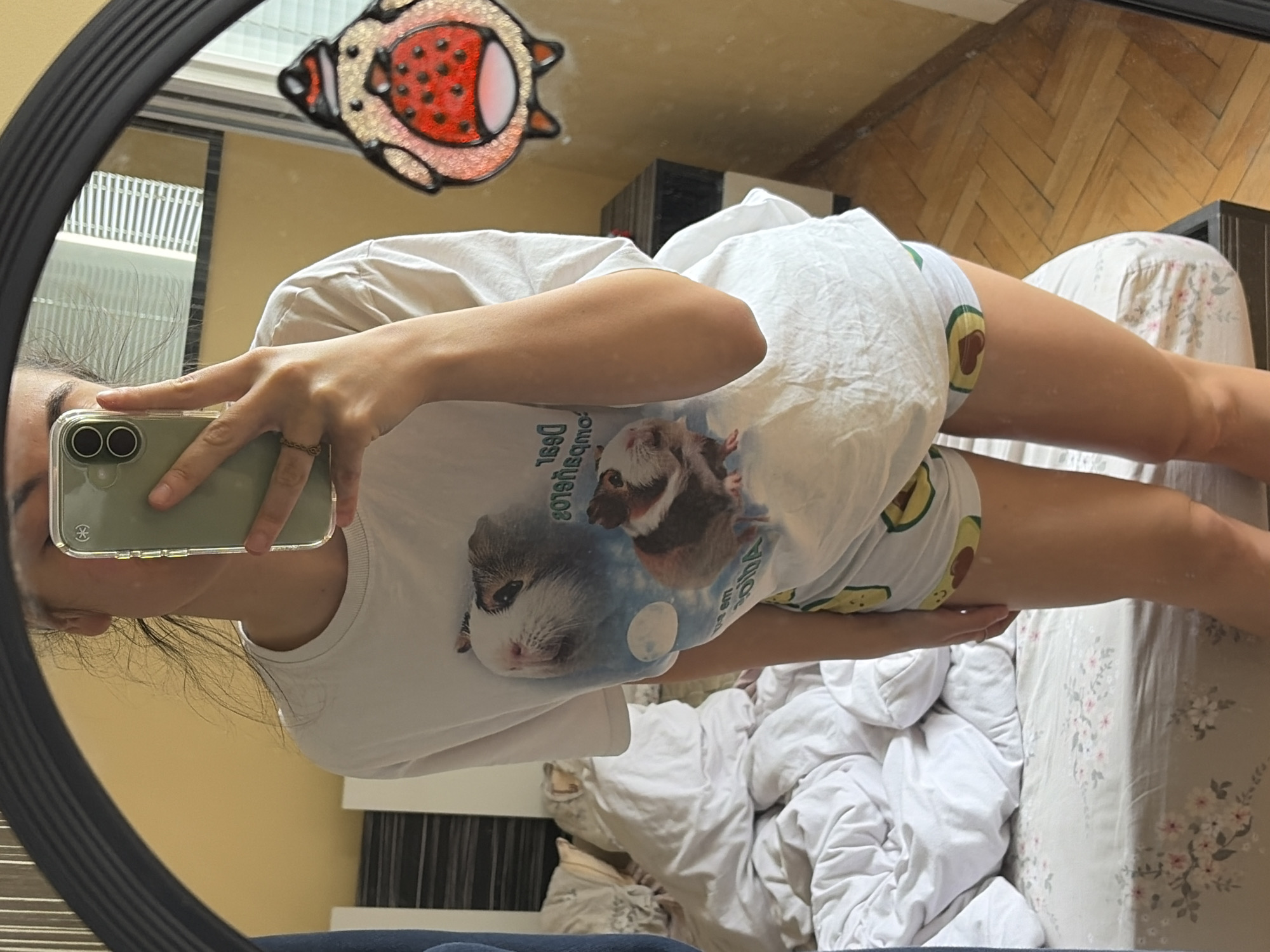 A young woman is taking a selfie in a mirror. She is wearing a white t-shirt with a graphic of guinea pigs and a pair of white shorts with avocado prints. The background shows a messy bed, a wooden floor, and a yellow wall. There is also a cute cartoon penguin sticker on the mirror.