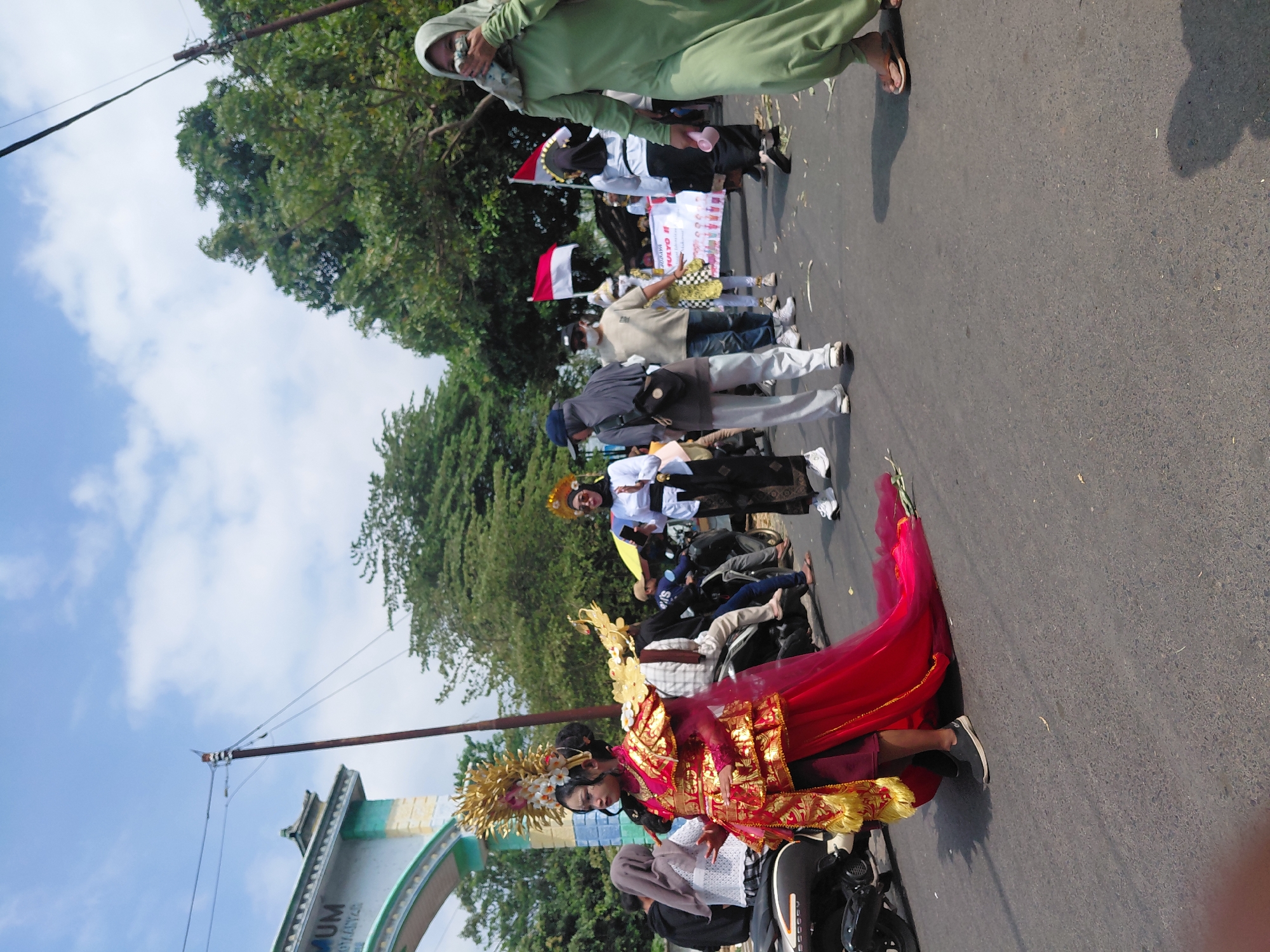 In Bimo, Indonesia, a street procession is underway under a bright, partly cloudy sky. A young person in the foreground is adorned in a striking traditional gold and red costume, complete with an elaborate headpiece. Their attire includes a gilded, tiered tunic, a deep red underskirt, and a flowing red train that trails onto the asphalt road. The person is wearing black sneakers.
Behind this central figure, other participants are visible. One individual, to the right of the foreground figure, wears a white long-sleeved shirt, black pants, and a black headscarf, holding what appears to be a smartphone. Further back, a person in a light green long-sleeved dress and a matching headscarf is walking away from the camera, covering their mouth and nose with their hand.
Several people are walking in the background. Some are wearing masks. A banner with text, including "JAYO II", is held by some participants. Indonesian flags (red and white) are also displayed. Motorcycles are parked and being ridden by some individuals in the procession.
The environment includes a large archway in the background with inscriptions, possibly indicating an entrance or landmark. Lush green trees line the sides of the road, and utility poles with wires are present. The road surface is asphalt, and scattered debris, possibly dried leaves or paper, is visible on the ground. The scene suggests a cultural or festive event taking place in a public area.