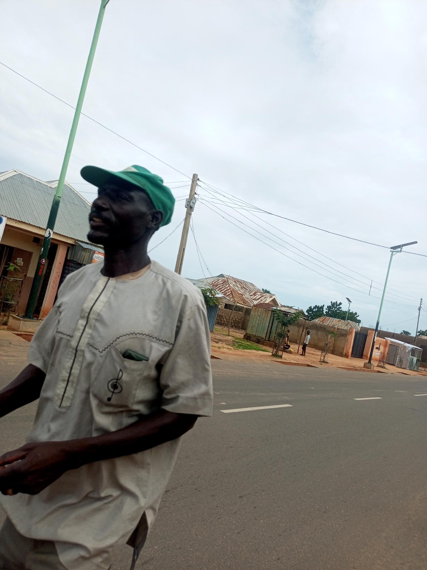 A low-angle, medium shot captures a dark-skinned man wearing a green baseball cap and a light-colored, collared shirt with decorative stitching and a treble clef emblem. He is looking to his left, with his mouth slightly open, and his right hand is visible in the lower left of the frame. He is standing on a paved road with white lane markings. In the background, a green pole with a light fixture rises vertically on the left, and another similar pole is visible in the mid-ground on the right. The scene depicts a street in Tempure, Nigeria, with simple buildings and corrugated metal structures lining the right side of the road. A few other individuals are sparsely visible in the distance. The sky is overcast with pale clouds.