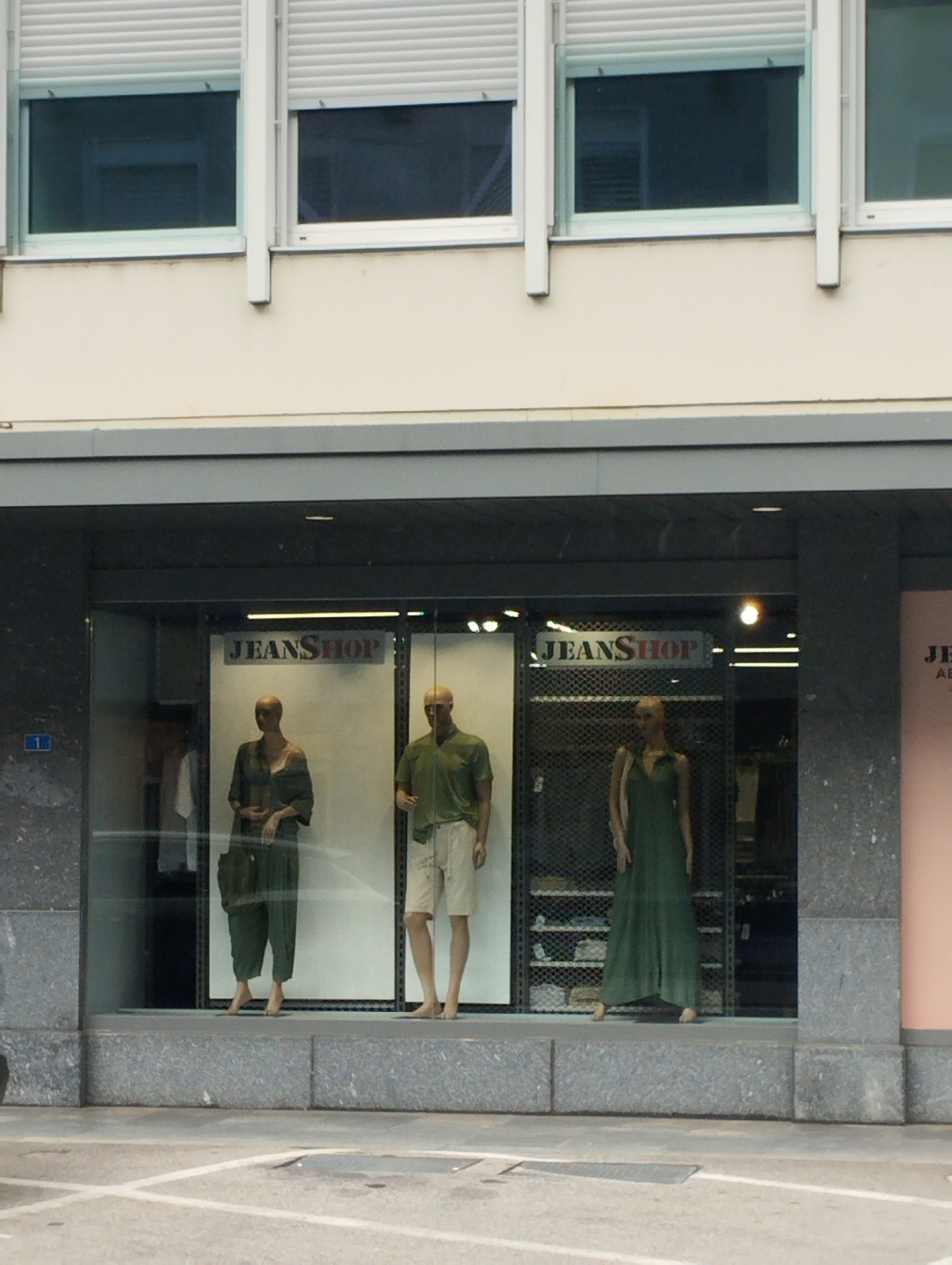 The image shows a storefront display window of a "JEANShop." In the window, there are three mannequins. The left mannequin is female, wearing a green jumpsuit and holding a dark green purse. The middle mannequin is male, wearing a green short-sleeved shirt and light-colored shorts. The right mannequin is female, wearing a long, flowing green dress. All three mannequins are barefoot. The storefront features a dark grey stone facade. Above the display window are three rectangular windows with white blinds pulled down. The background of the display window shows shelving units and more clothing.