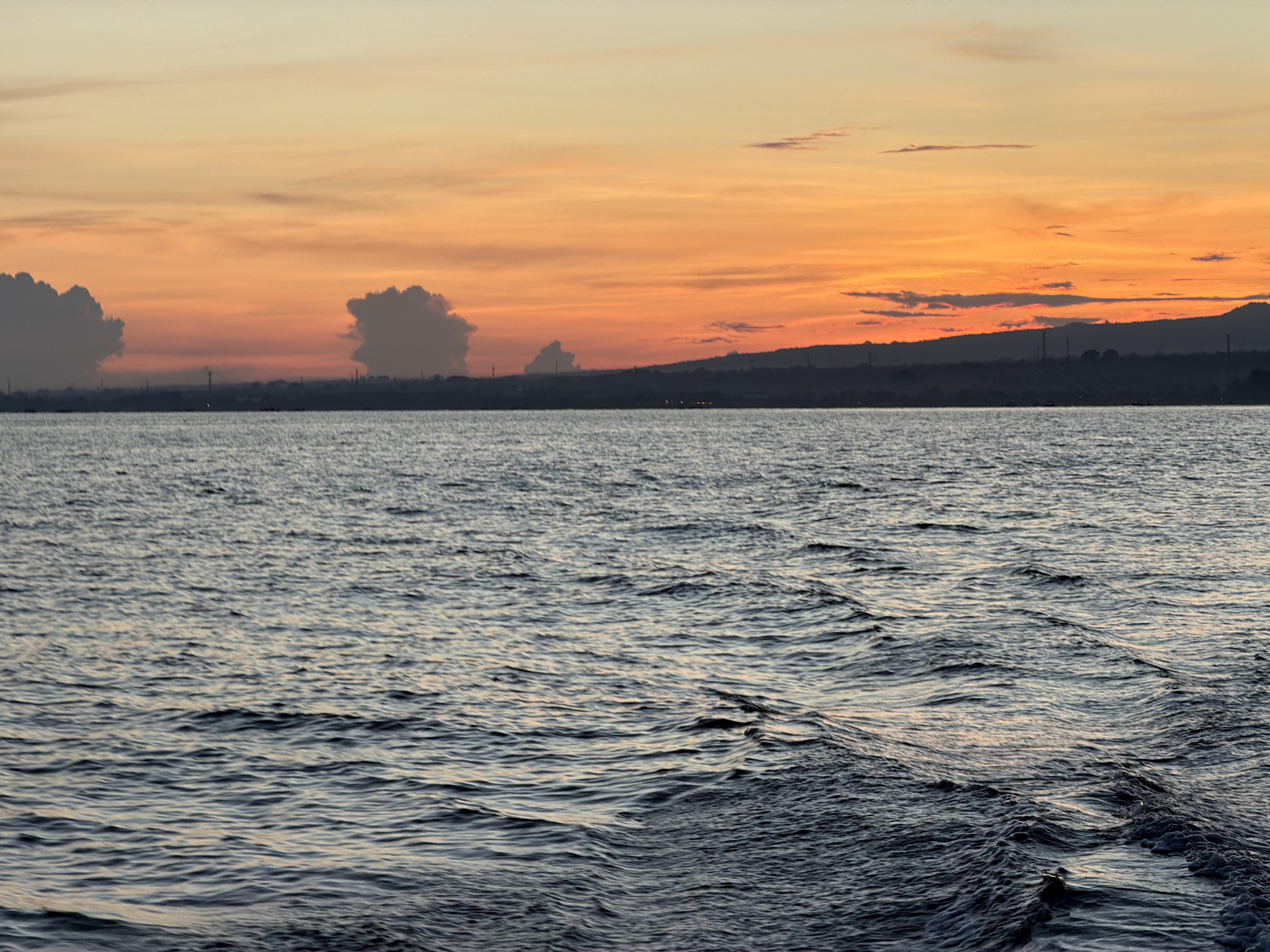 The image depicts a body of water under an orange and yellow sunset sky with mountains and what appear to be industrial structures along the distant shoreline. The water is choppy with visible wave patterns, likely indicating movement or wind. The shoreline silhouette includes hills or mountains, power lines, and possible buildings or towers. It is likely photographed near Kaliasem, Indonesia, based on the prompt.