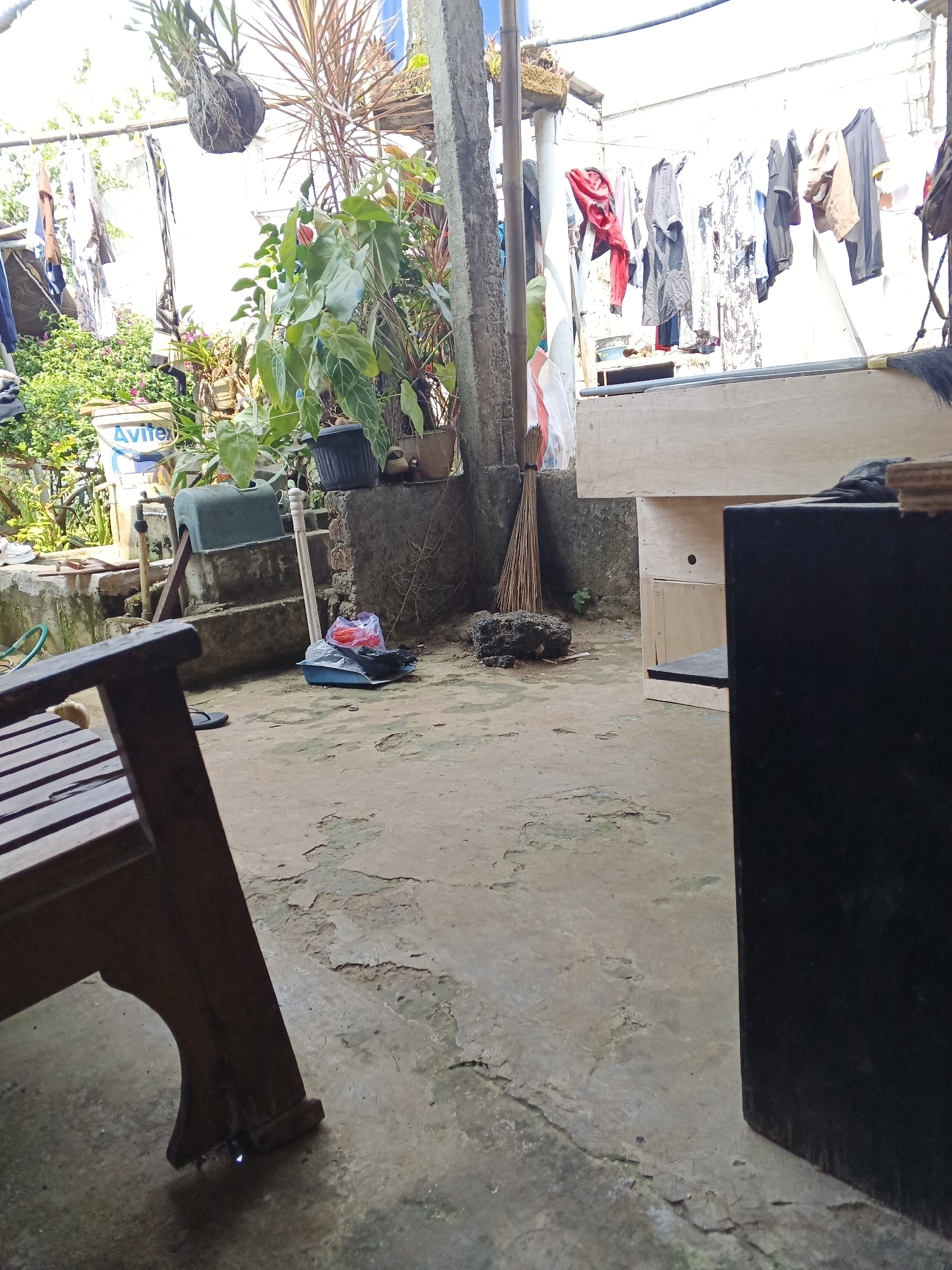 The image depicts an outdoor backyard area, likely in Warungkiara, Indonesia, characterized by a mix of functional and domestic elements. The ground is paved with a cracked, weathered concrete surface. To the left, a wooden chair with a slatted seat and backrest is positioned prominently in the foreground. Adjacent to the chair, a blue bucket labeled "Avitex" is visible, alongside other containers and plants. In the background, a clothesline holds several items of laundry, including shirts and trousers, drying in the open air. Various plants, some in pots and others growing wild, add greenery to the scene. A broom stands upright next to a concrete structure. Further back, a large wooden cabinet or workbench is partially visible. The overall impression is of a lived-in, working space, possibly a residential backyard.
