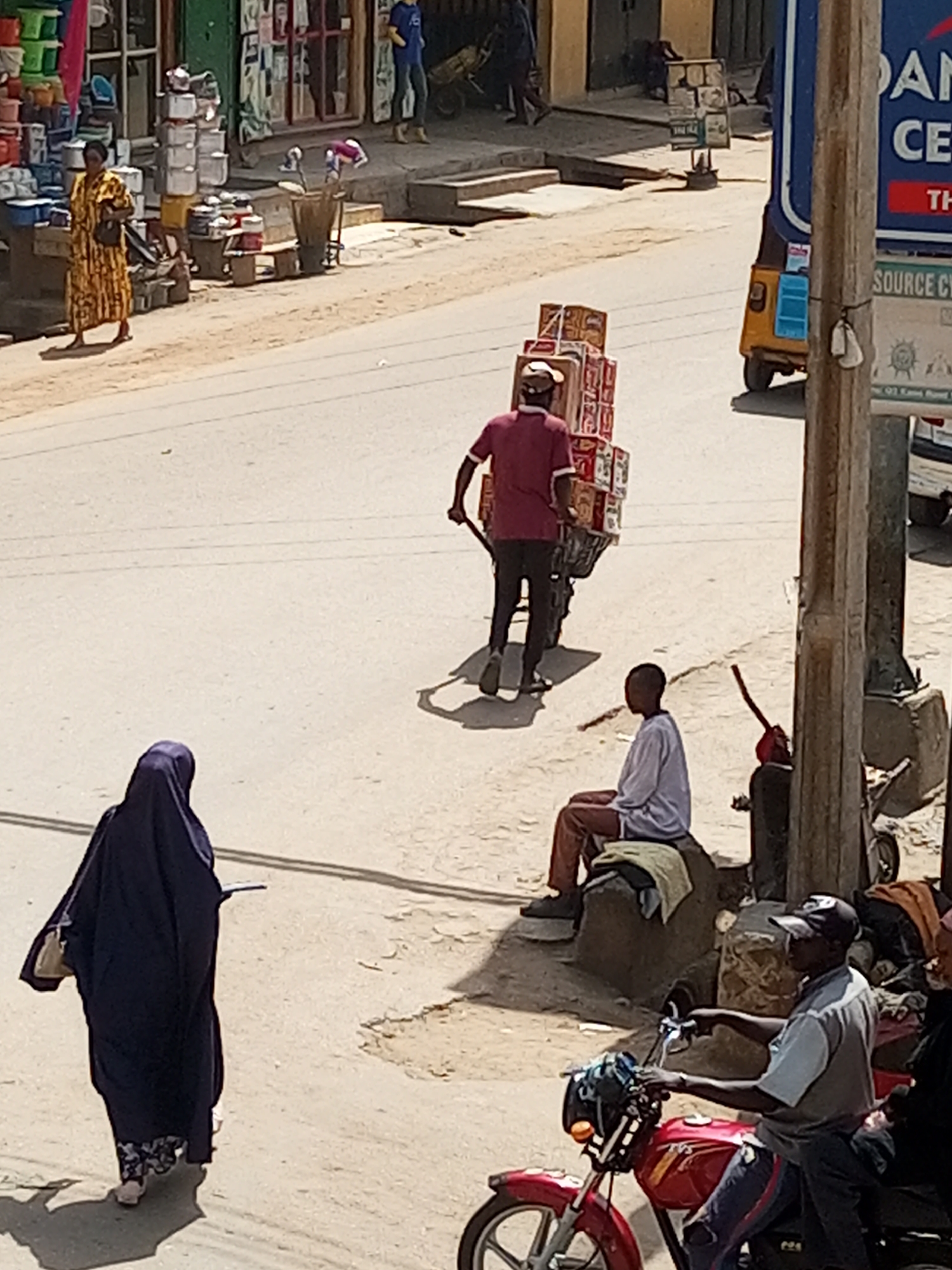 This image captures a bustling street scene in Maiburiji, Nigeria, under bright daylight, likely midday, indicated by the strong shadows. The setting appears to be a commercial or market area with shops lining one side of a road, which transitions from unpaved dirt to a smoother surface.
Several individuals are visible. In the foreground-left, a person wearing a dark blue abaya or hijab with a floral pattern at the hem walks away from the viewer, carrying a light-colored shoulder bag and holding what appears to be a book or tablet. On the foreground-right, a man wearing a grey shirt, dark pants, and a black baseball cap sits astride a red motorcycle with "TVS" visible on its tank, holding the handlebars.
In the mid-ground, a man in a red shirt and a light cap pushes a two-wheeled handcart heavily loaded with stacked cardboard boxes, suggesting commercial delivery or transportation of goods. Near him, towards the right, another person sits on a large stone or concrete block, wearing a light long-sleeved shirt and brown trousers. Further down the street on the left, a woman in a patterned yellow dress walks along the roadside, holding a bag, amidst shop displays of various household items like stacked pots and buckets.
The background features more shops and blurry figures. A yellow auto-rickshaw (keke napep) is partially visible on the right. A prominent blue and white sign on the right side of the road is partially obscured by a wooden pole, showing fragmented text like "AN", "CE", "TH", and "SOURCE CY". Below it, the text "Q3 Kaus Road" can be discerned. The scene depicts everyday life and commerce in a vibrant street environment.