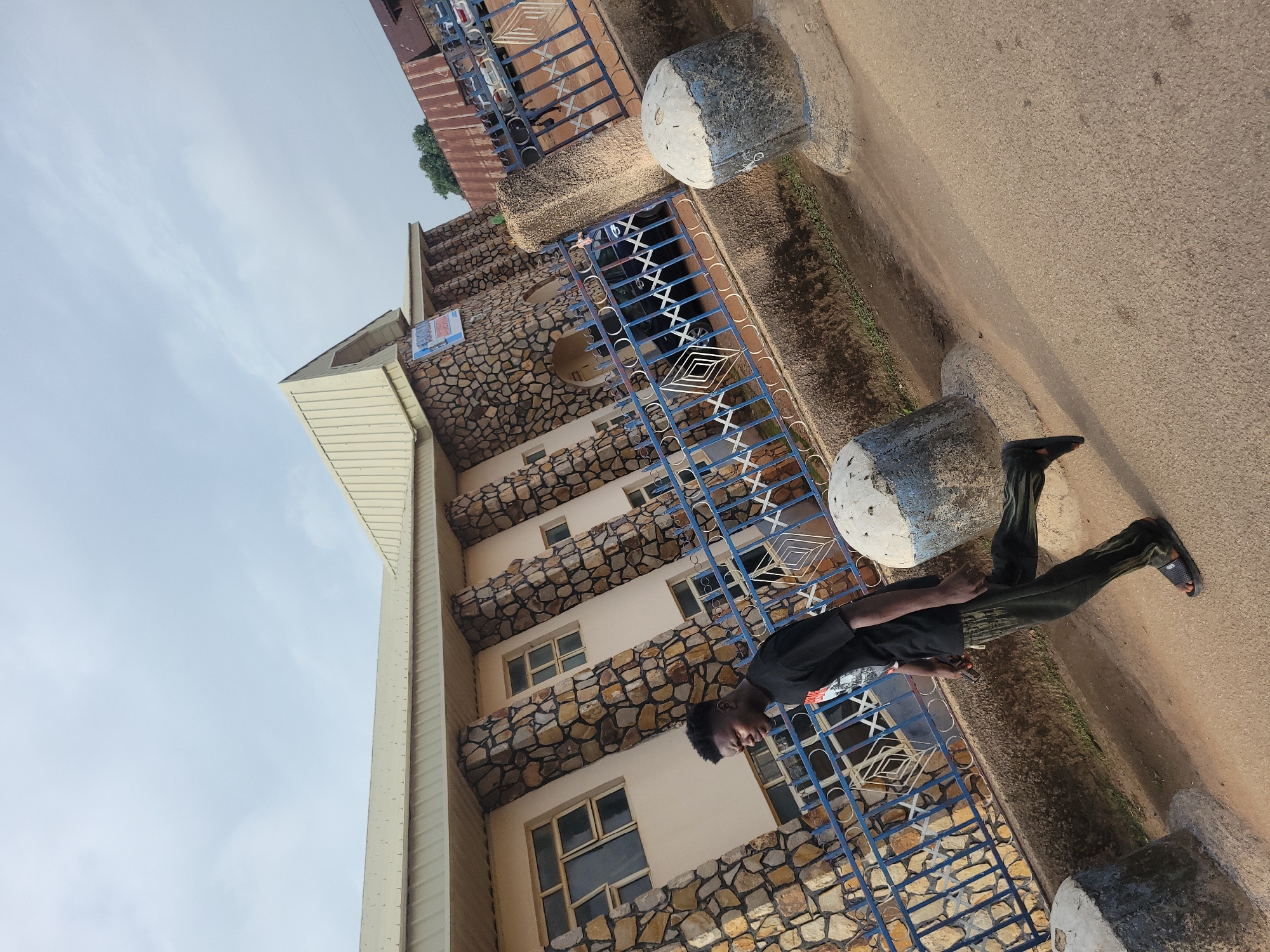 A young Black male walks along a paved path in front of a building in Jos, Nigeria. He is wearing a black t-shirt and dark pants with a striped pattern, and black sandals. The building has a cream-colored facade with stone accents and a beige, sloped roof. A blue metal fence with diamond patterns runs along the front of the building. Two large, cylindrical concrete bollards, painted with blue and white accents, are visible in the foreground on the right and left of the path. The sky is overcast.