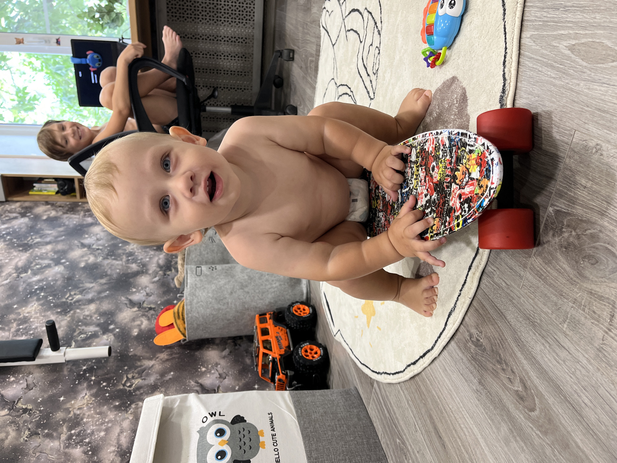 The image depicts a close-up of a baby sitting on a colorful toy skateboard on a round rug. The baby is wearing a diaper and has blonde hair and blue eyes. In the background, a slightly older child is sitting in a chair at a desk with a laptop, smiling. The room appears to be a child's play area or bedroom, with toys and storage bins visible. The wall has a dark, starry pattern. The overall scene suggests a domestic setting, likely a home. There is no information in the image to suggest this location is in Nizhny Novgorod, Russia.