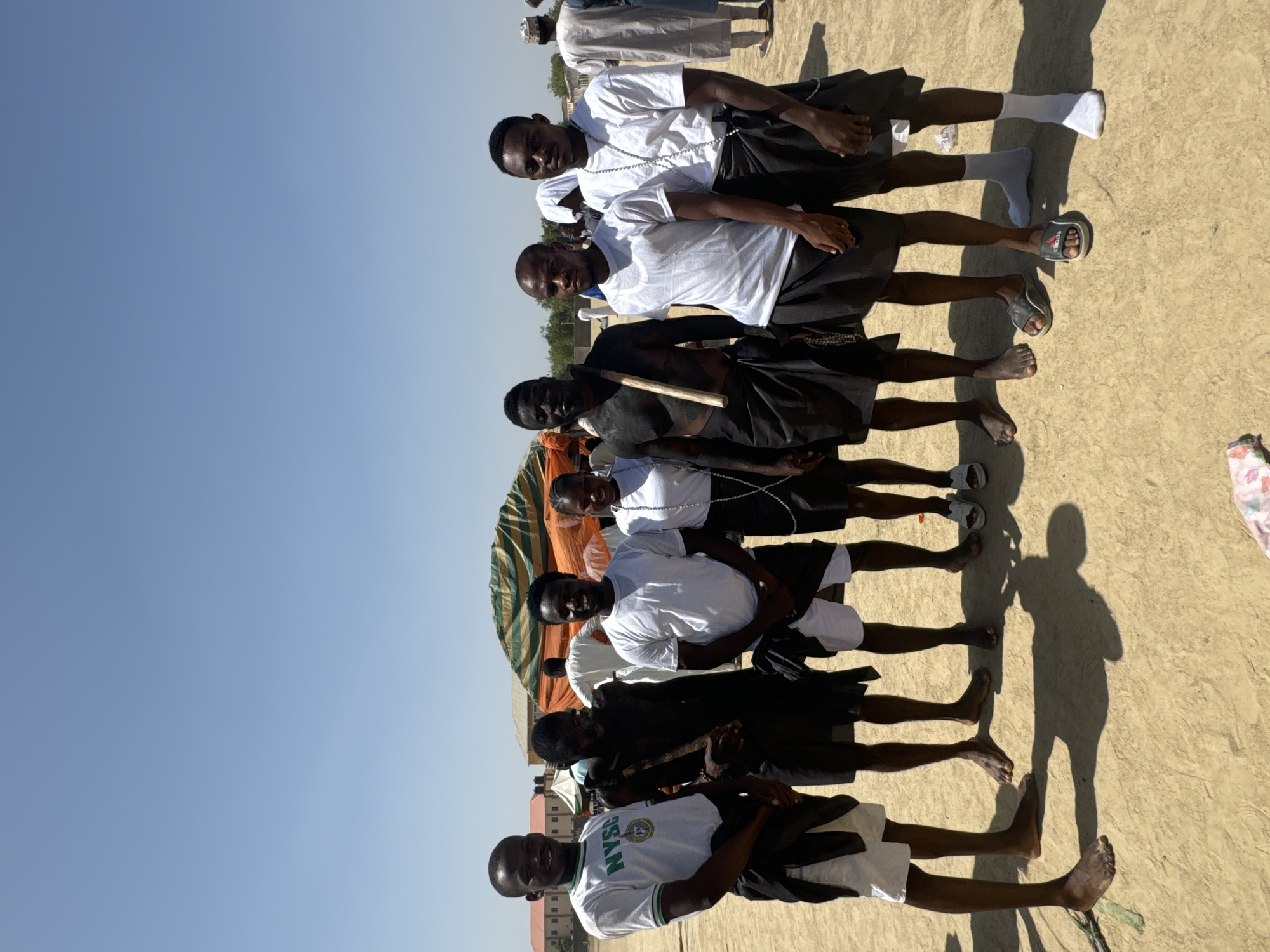 A group of seven young men stand close together outdoors under a clear blue sky. The location is presumed to be in Maiduguri, Nigeria, due to one of the men wearing a shirt with "NYSC" on it, referring to the Nigerian National Youth Service Corps.
The men appear to be in their late teens or early twenties, are dark-skinned, and have short black hair. They are all wearing shorts or trousers that are black, brown, or white. The top-most men wear white shirts, except for the front-most man, who wears a NYSC shirt. Some of the men wear white beaded necklaces.
The ground is beige. A dark-skinned man has dark pigments spread across his face. A green and brown striped awning is visible in the background. The men appear to be posing.