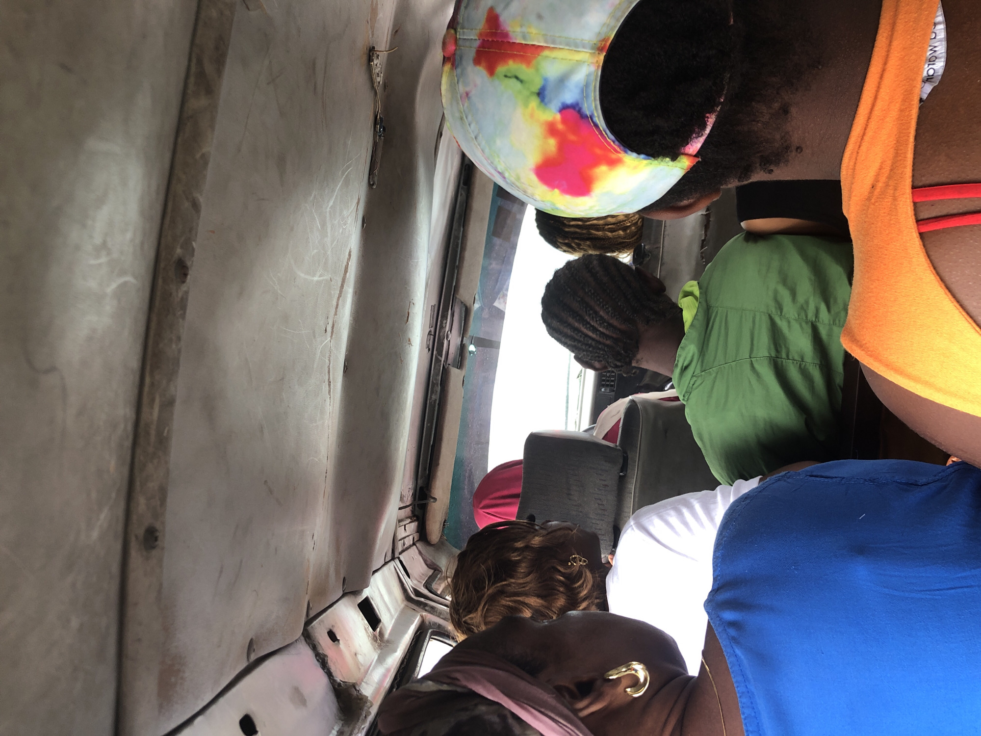 The image depicts the interior of a crowded vehicle, likely a bus or shared taxi, in Lekki, Nigeria. The perspective is from within the vehicle, looking forward. The ceiling of the vehicle is visible and appears worn, with exposed wires and some damage. Several people are seated inside, mostly seen from behind or the side.
In the foreground, on the left, a person wearing a blue top and a dark headscarf with a gold earring is partially visible. To their right, another person with reddish-brown curly hair and a white t-shirt is seated. Further back, a person in a bright green shirt is visible, their head tilted forward. To their right, a person wearing a colorful tie-dye baseball cap is seen from behind. In the middle background, a person wearing a pink headscarf is seated. The overall environment suggests a mode of public transportation, likely in an urban setting in Lekki, Nigeria, given the context. The lighting appears to be natural daylight, filtering in from outside.