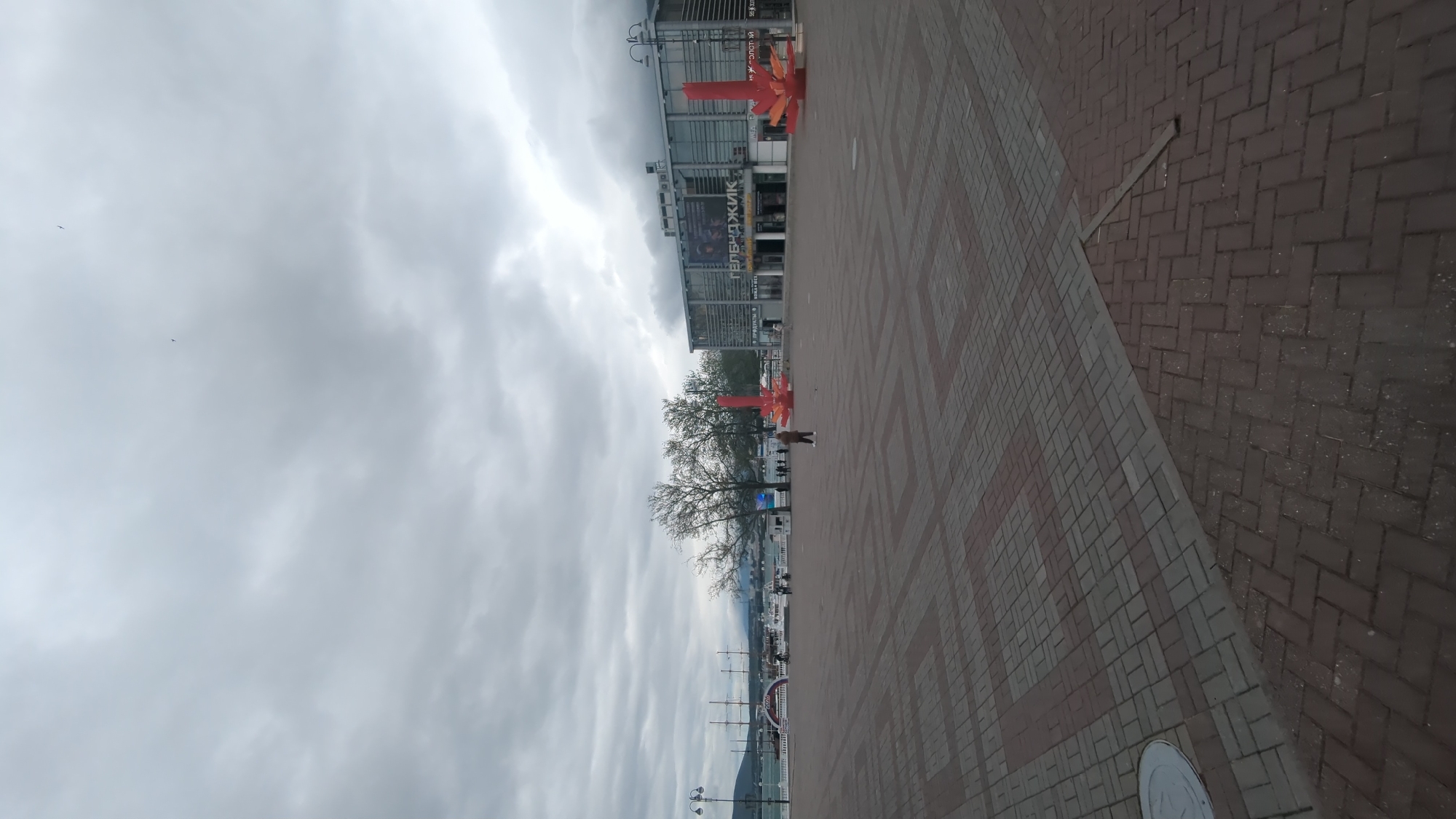 The image shows a partially cloudy sky over a brick-paved walkway in Gelendzhik, Russia. To the left of the walkway are a few trees, a building with lettering in Russian, and some red decorations. A person is seen walking near the trees. In the background, masts of ships or sailboats are visible along the coastline.