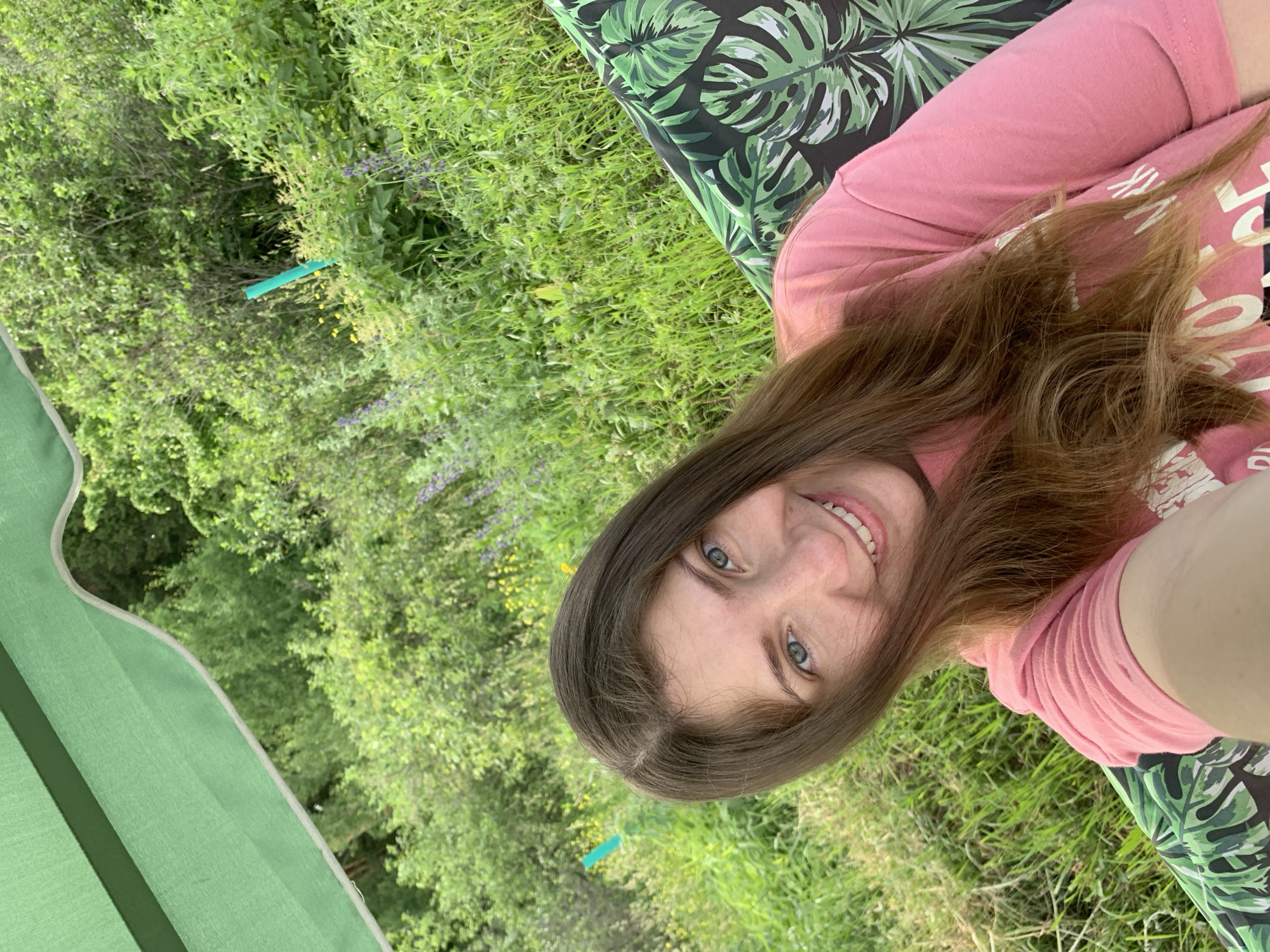 A woman with long brown hair and blue eyes smiles at the camera. She is wearing a pink t-shirt and sitting on a swing set with a green and black palm leaf patterned cushion. The swing set is outdoors, surrounded by lush green foliage and trees. A bright green pole is visible in the background. The image appears to have been taken on a sunny day. The setting suggests a park or a backyard in a natural, possibly rural, environment within or near Chaikovsky, Russia.