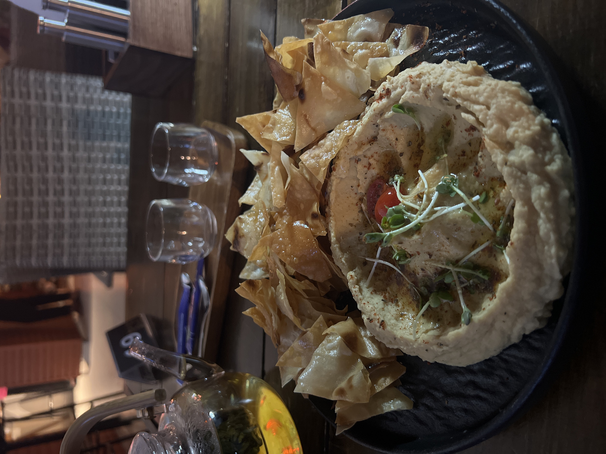 The image depicts a plate of hummus with pita chips, likely served in a restaurant. Two small, empty glasses and a teapot are visible to the left of the plate. The setting appears to be indoors on a wooden table. Given the prompt information, this scene is likely in Bac Nha Trang Ward, Vietnam.