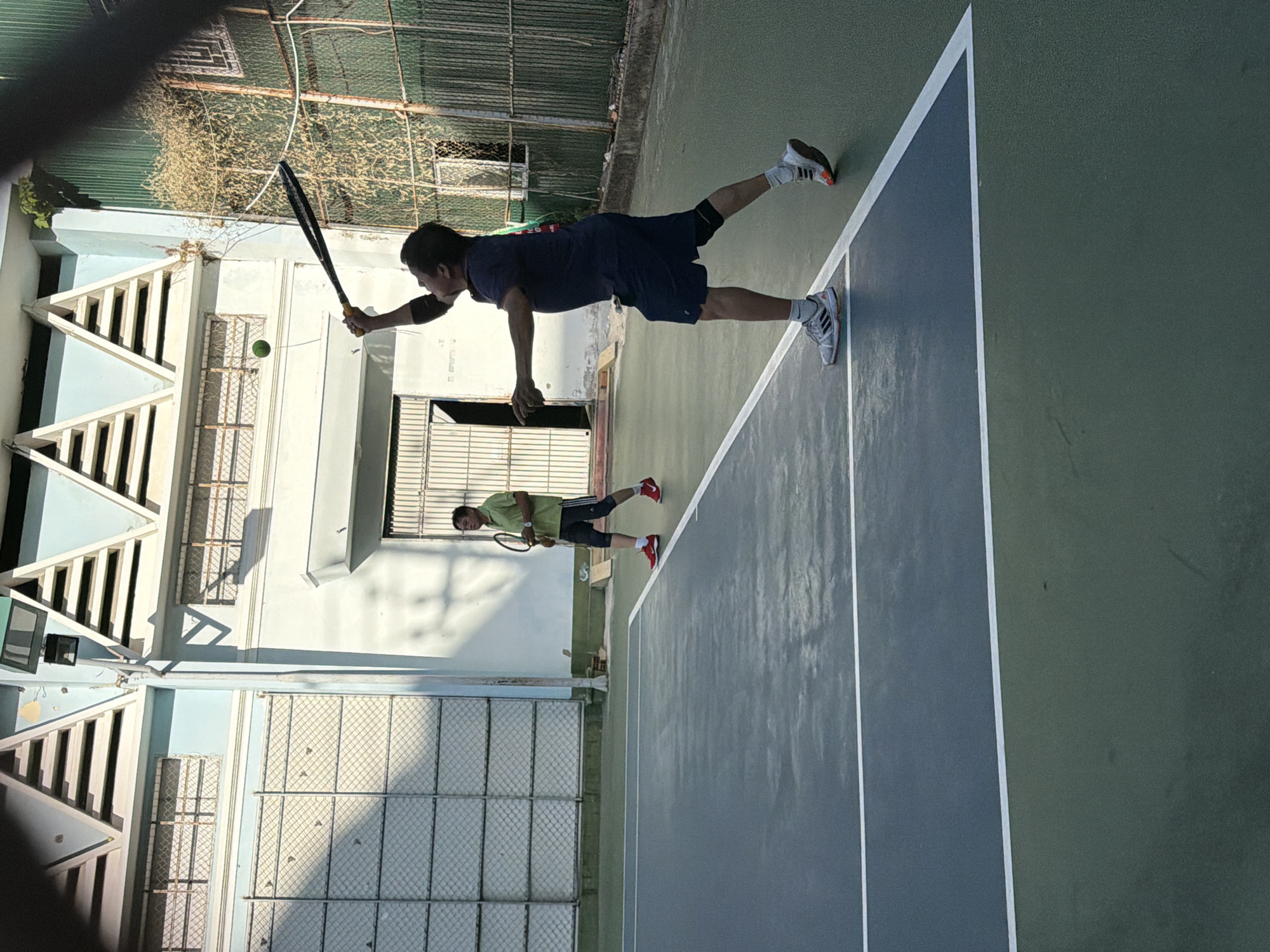 Two men are playing tennis on an outdoor court in Nha Trang, Vietnam. The man in the foreground, wearing a dark blue t-shirt and shorts, is mid-swing, hitting a tennis ball that is visible in the air above him. He is wearing athletic shoes and a knee brace. The man in the background, wearing a green t-shirt and dark shorts, is holding a tennis racket and looking towards the player in the foreground. The tennis court has a blue and green surface with white lines. In the background, there is a light blue building with white triangular architectural features and windows with metal grates. A green metal fence borders the court and the building. The image is taken from a slightly elevated, angled perspective.