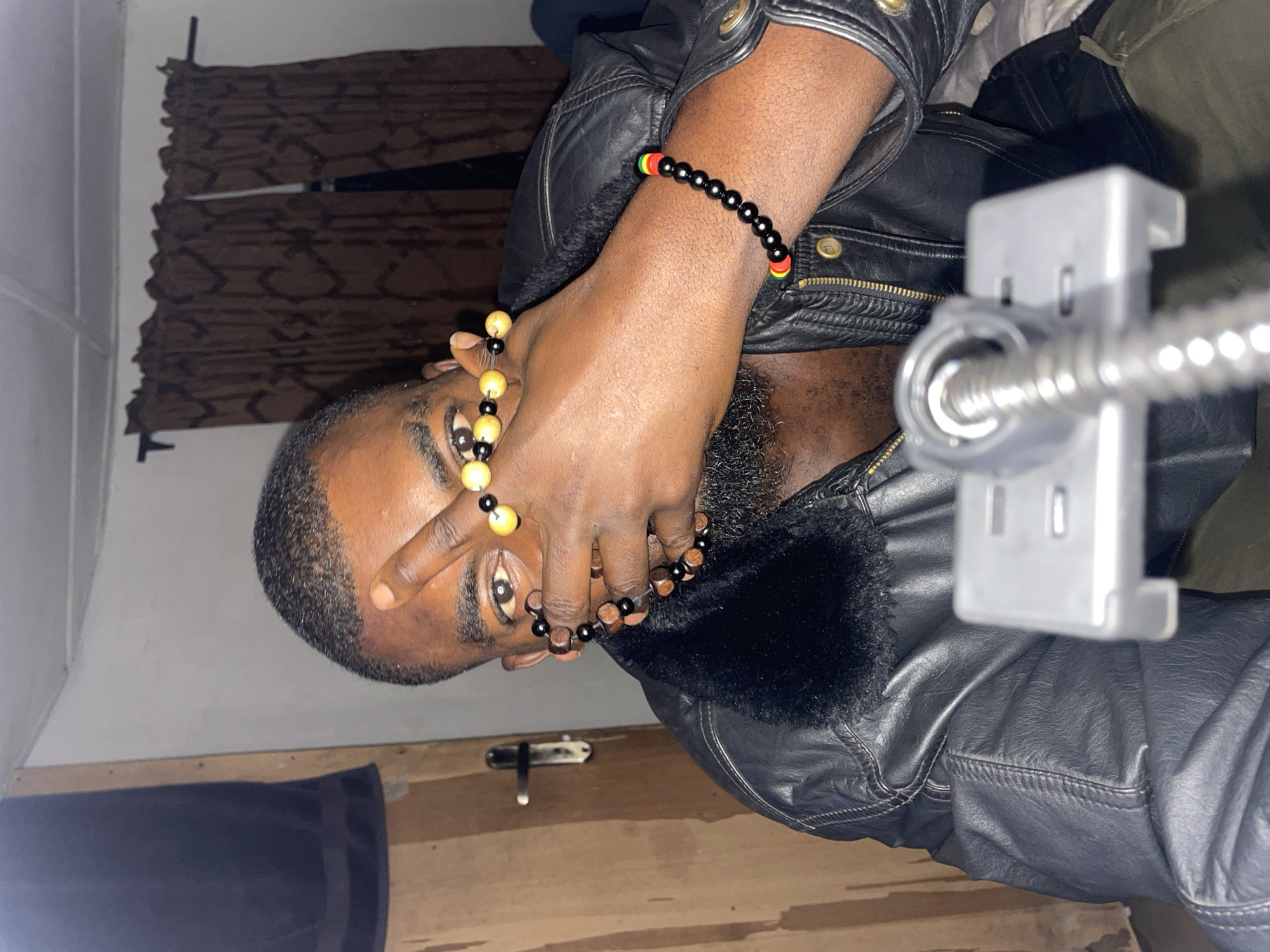 A medium shot of a man in an indoor setting. The man is dark-skinned, with short dark hair, a beard, and wearing a black leather jacket with a fur collar. He is holding his hand up near his face, adorned with beaded bracelets. The man is looking directly at the camera. Behind him are neutral-colored curtains. The location is unknown, and there is no evidence to suggest this photograph was taken in Jos, Nigeria.