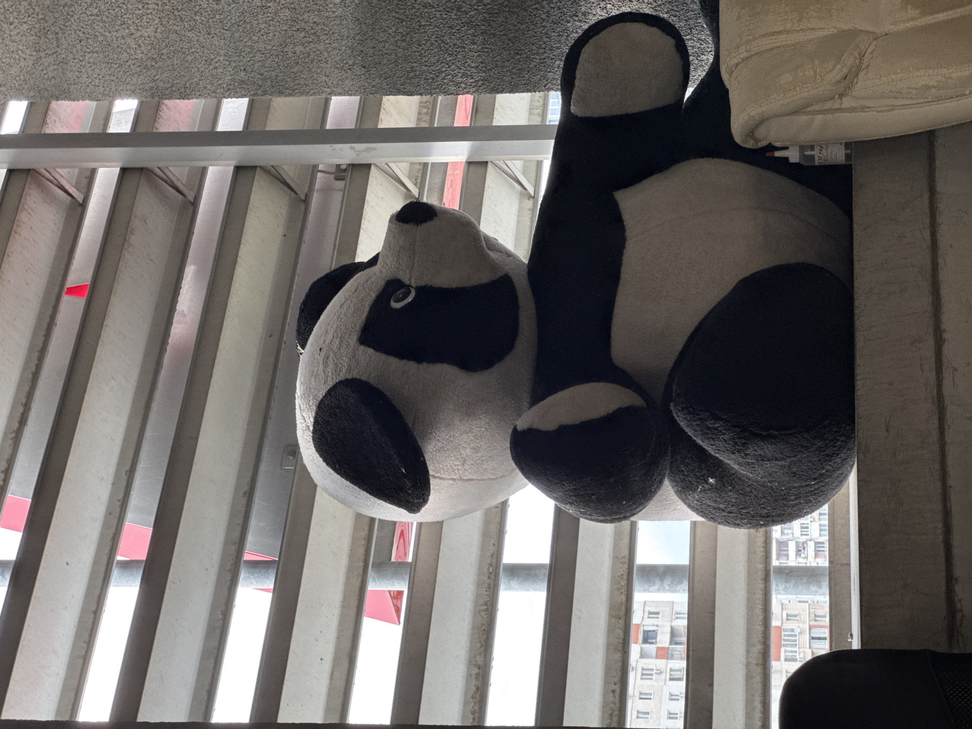 A close-up, upside-down shot captures a plush panda toy draped over a railing. The panda's head is on the left, with its distinctive black eye patches and a black nose. Its body, made of white and black fur, extends to the right, with its hind legs also visible.
Behind the panda, a series of vertical, light-colored metal slats create a striped pattern. Gaps between these slats reveal a bright, overcast sky. In the distance, through the slats, is a view of urban buildings with multiple windows. The architecture suggests a contemporary city setting, consistent with Sofia, Bulgaria. A small, clear bottle with a red cap and some text in a non-Latin script is partially visible underneath the panda's right hind leg, nestled between it and a beige, textured object that appears to be part of a couch or cushion. The overall lighting is bright, indicating daytime. The image is a still shot, with no discernible activity or emotional cues.