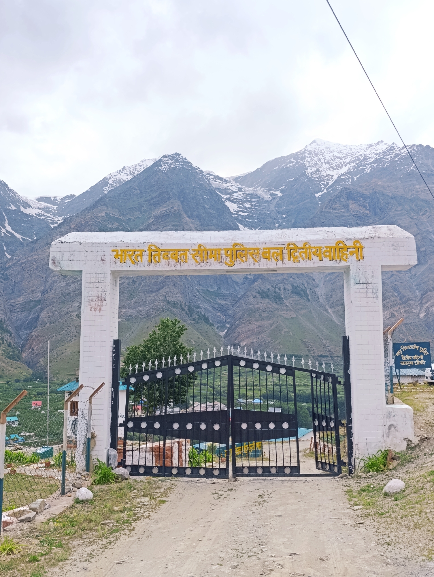 The image depicts the entrance gate to the "Bharat Tibet Seema Police Bal Dwitiya Vahini" in Tandi, India. The gate is made of white brick pillars and a white archway inscribed with yellow text. A large black metal gate with decorative circles stands open. Behind the gate, a green tree is visible, and beyond that, the slopes of snow-capped mountains dominate the background. The foreground shows a dirt road leading to the gate, with sparse grass and some rocks. A barbed wire fence runs along the left side of the entrance. A sign with Hindi text is visible on the right side of the gate. The sky is overcast.