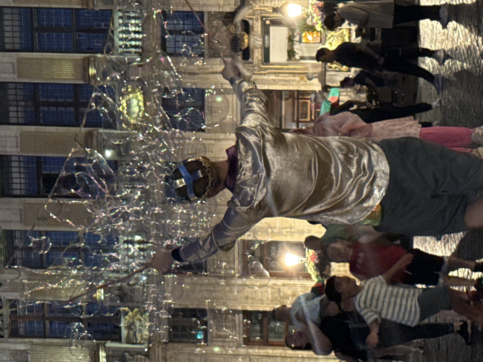 The image depicts a lively scene on the Grand Place in Brussels, Belgium, at night. A performer, facing away from the camera, is creating a cascade of large, iridescent soap bubbles using wands. They are wearing a shiny, champagne-colored tunic and a blue and gold crown-like headpiece. The bubbles catch the light, creating a magical effect.
In the foreground, a diverse crowd of people are gathered, watching the spectacle. To the left, a woman holds a child, both looking up at the bubbles. Other individuals, including two young boys, are visible in the mid-ground, their attention also directed towards the performer. One boy is wearing a striped shirt and shorts, while the other is in a red t-shirt and dark shorts, appearing to dance or move with excitement.
The background features the iconic architecture of the Grand Place, with its ornate guildhalls and buildings illuminated by warm, ambient light. Streetlights and illuminated windows are visible, casting reflections on the wet cobblestone ground, suggesting it may have recently rained. The overall atmosphere is one of festive celebration and enchantment.