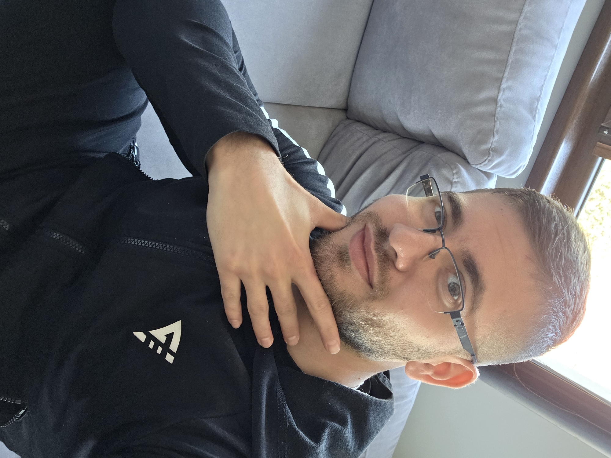 A man is lying on a gray couch. He is wearing glasses and a black jacket with a white logo. His hand is on his neck. A window with light streaming in can be seen next to the couch. No information in the image confirms the location to be Yagodovo, Bulgaria.