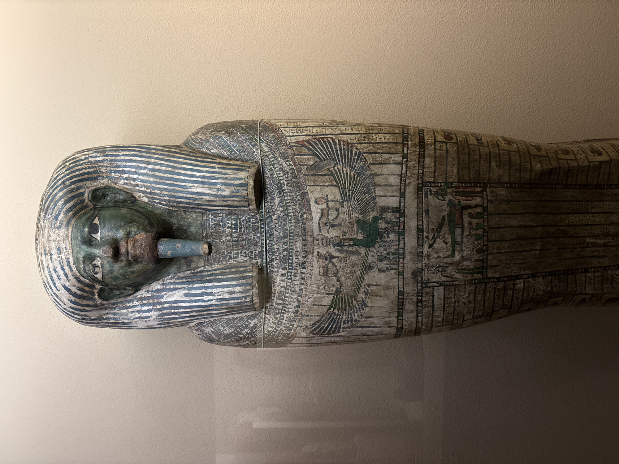 The image shows an Egyptian sarcophagus, likely on display at the Oriental Institute Museum in Chicago, United States. The sarcophagus is painted and decorated with traditional Egyptian symbols and motifs. It features a human-shaped face with a funerary mask and a full-body depiction with hieroglyphic inscriptions and images of deities and winged creatures. The sarcophagus is positioned horizontally, with a plain, light-colored wall in the background. The surface of the sarcophagus shows signs of age and wear. It is encased in glass.