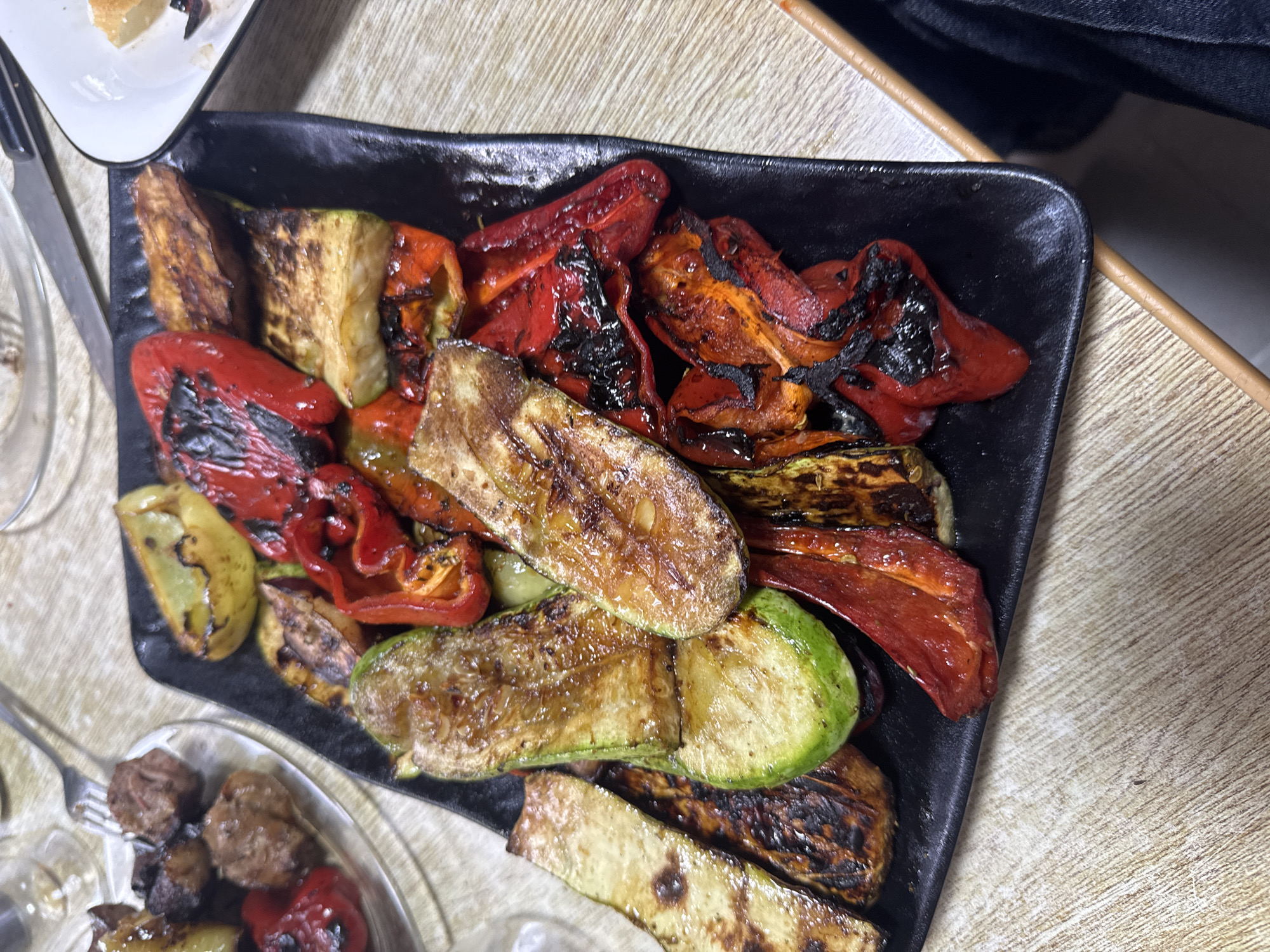 A close-up, overhead shot shows a black rectangular plate filled with grilled vegetables, including zucchini, bell peppers, and eggplant. The vegetables are charred in places, indicating they were cooked over an open flame. The plate is placed on a wooden table, with a small white dish containing a piece of food in the upper right corner and a glass plate with meat in the upper left. The scene appears to be a meal in Bishkek, Kyrgyzstan.