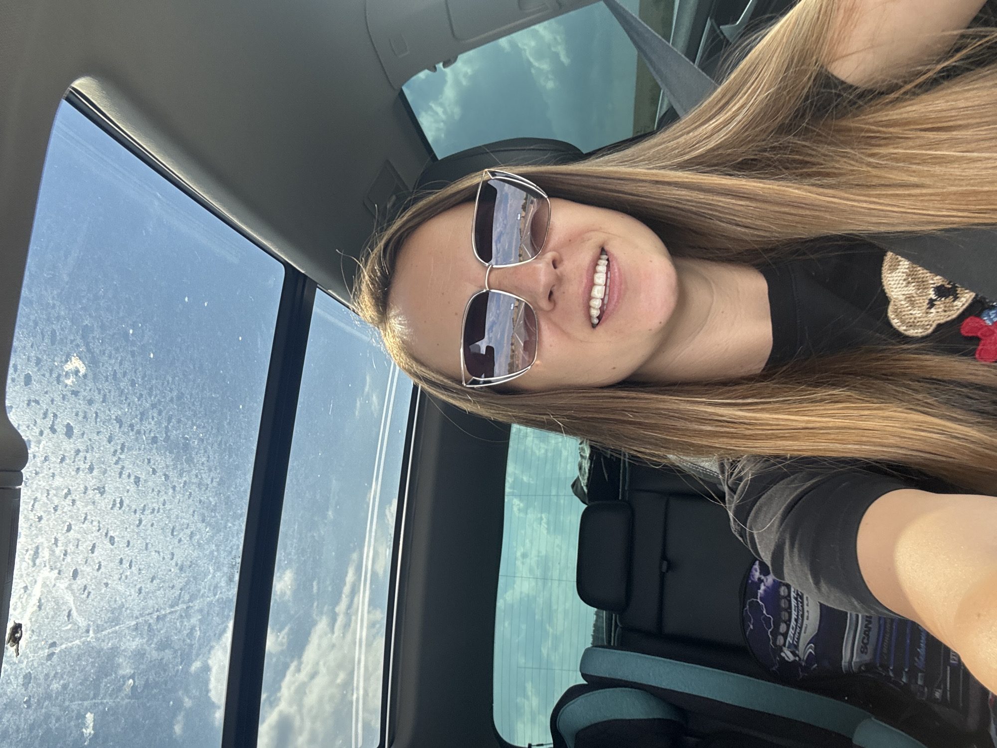 A woman with long, blonde hair and sunglasses is taking a selfie in a car. She is wearing a black t-shirt with a teddy bear graphic. The car has a sunroof, and the sky is visible through it. The back seat of the car contains a child seat and a bag with "SCANIA" written on it. The reflection in the woman's sunglasses shows a road and a landscape. The car appears to be in motion, possibly on a highway. The setting appears to be rural, with a wide open sky. The image was taken in Kozare, Bulgaria.
