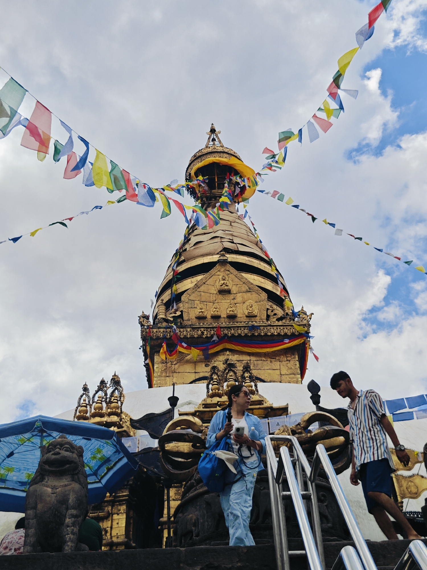 In Kathmandu, Nepal, a golden stupa, adorned with prayer flags fluttering against a cloudy sky, dominates the scene. In the foreground, two people ascend a set of stairs. On the left, a woman in a light blue outfit holds a phone and a water bottle, carrying a blue bag. To her right, a young man in a striped shirt and blue shorts walks down, looking at the stairs. Behind them, a stone lion statue sits beside a blue umbrella, with more ornate structures of the stupa visible. The overall atmosphere suggests a pilgrimage or a visit to a significant religious site.