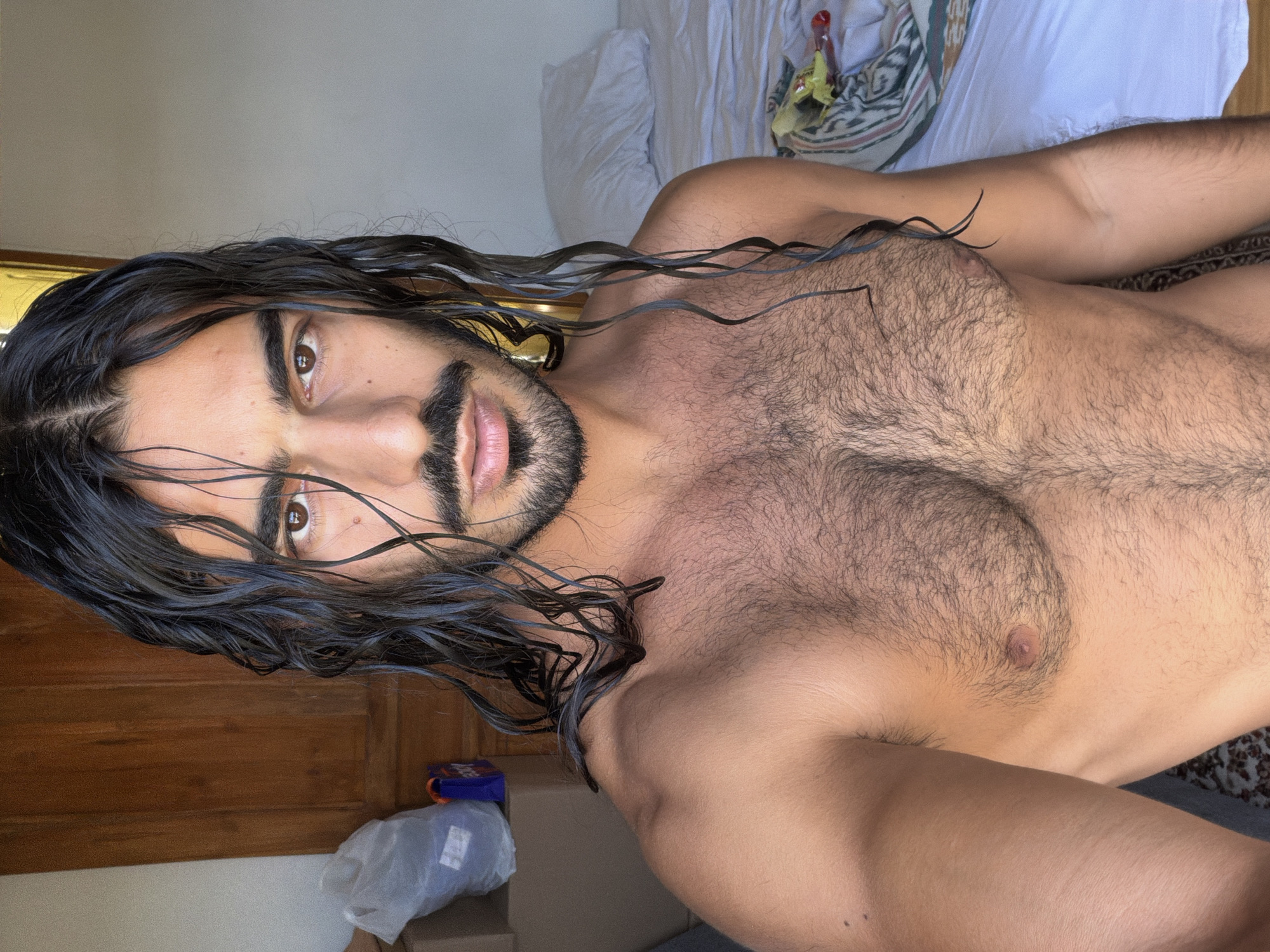A shirtless person with long, wet, dark hair and a beard looks at the camera. They have brown eyes and a mole below their right eye. The person's chest is covered in dark hair. To the left of the person, there are cardboard boxes and a white plastic bag. Behind the person is a bed with white sheets and a pillow, and a wooden door. The scene appears to be indoors. The location is in the city of North Kuta, Indonesia.