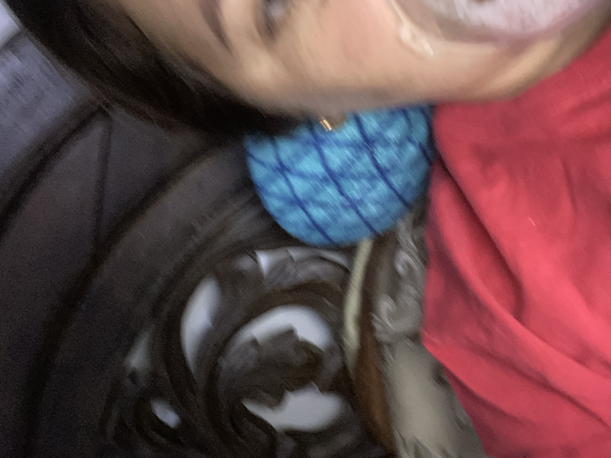 The media file is a close-up, blurry, and abstract image that appears to be a selfie taken by a person. The primary subject is a person's face, partially obscured and out of focus. A portion of their cheek, eye, and mouth is visible. They are wearing a gold earring.
Below the face, a bright blue object with a diamond-patterned texture is visible, likely a piece of clothing or an accessory. To the right, a large expanse of red fabric dominates the frame, also appearing to be clothing. The background is dark and indistinct, with some blurry shapes that suggest an indoor setting, possibly furniture or a dark container.
The image has a low resolution and is motion-blurred, making it difficult to discern specific details or activities. The lighting is dim. There is no visible text in the image. The overall impression is one of intimacy and informality, as is common with selfies, but the lack of focus and clarity prevents any definitive interpretation of the person's emotions or the context of the shot.